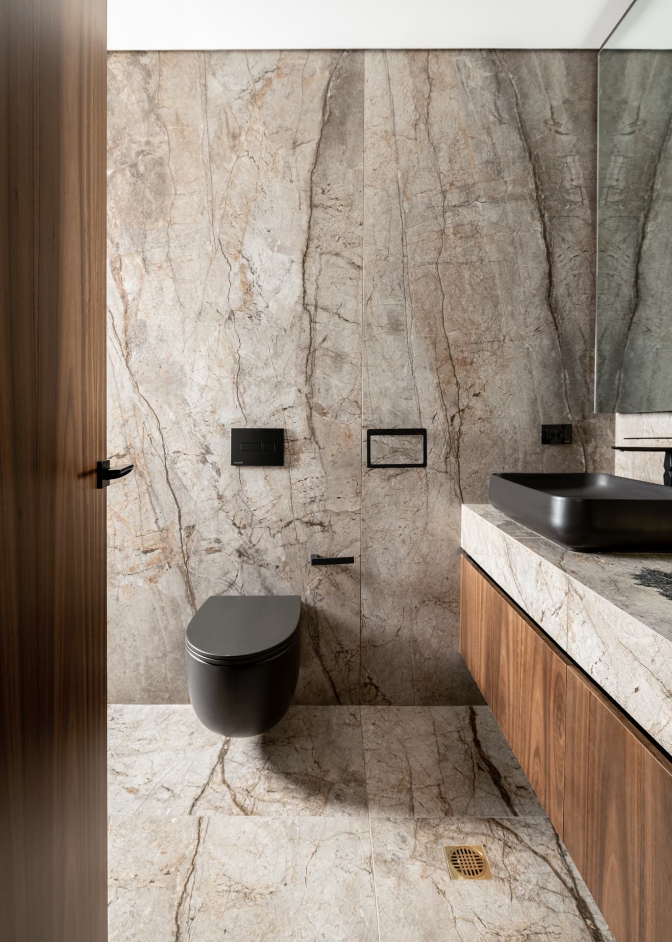 Wall-hung matte black toilet against floor-to-ceiling veined stone, beside a walnut vanity, in a bathroom of Argamán