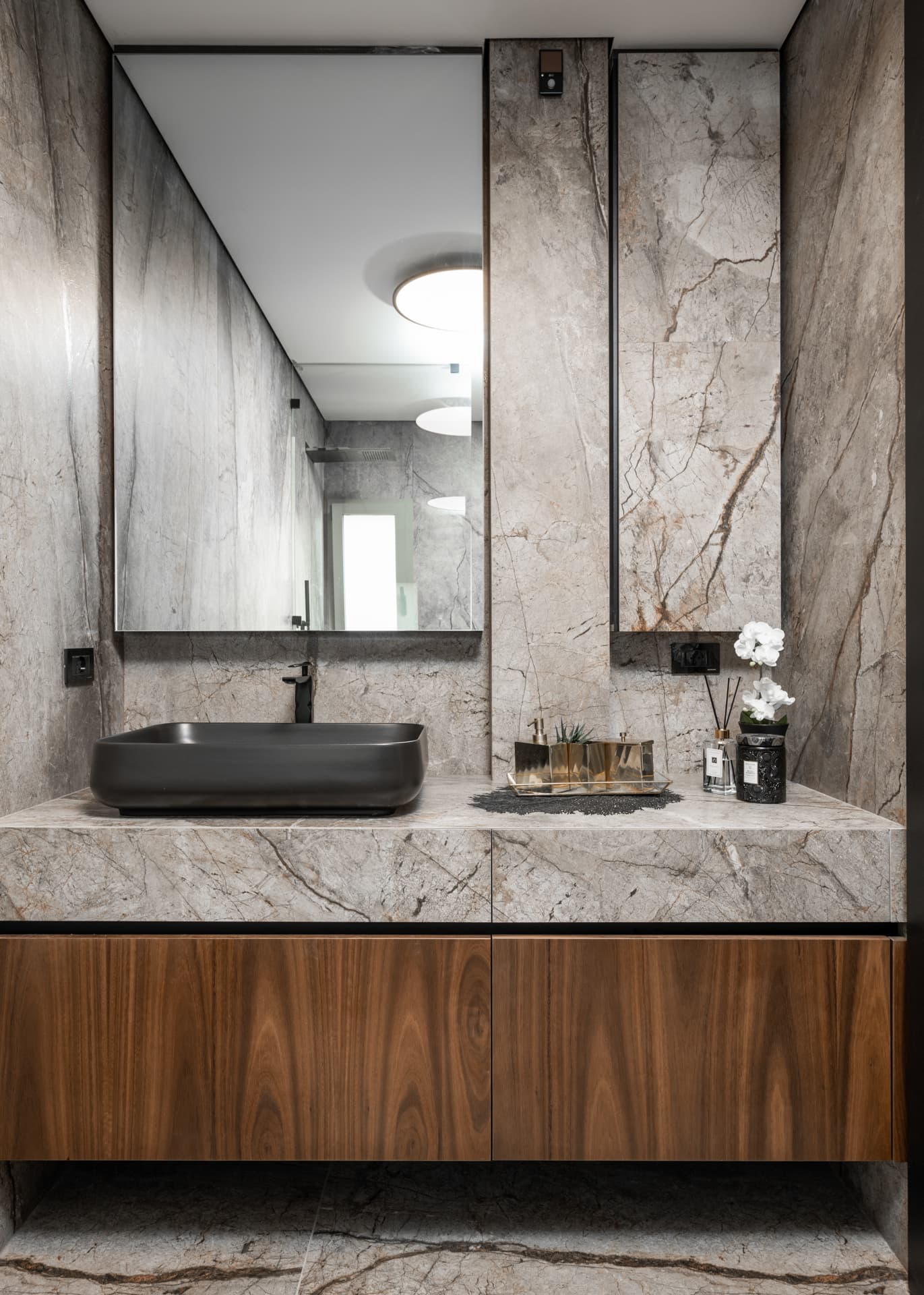 Floating walnut vanity with a black vessel sink on a veined stone counter under a slim steel-framed mirror in a bathroom of Argamán