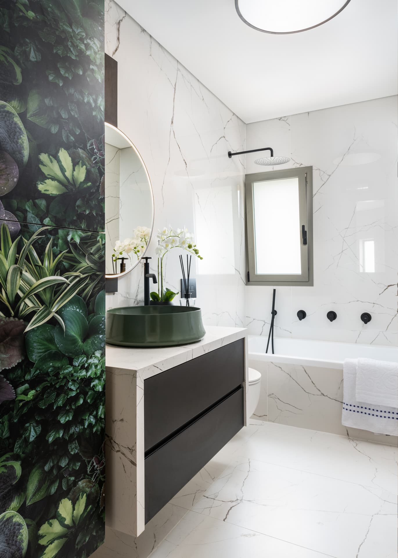 White veined-stone bathroom with a green vessel basin, a foliage-print wall panel and a bathtub with black fixtures in Argamán