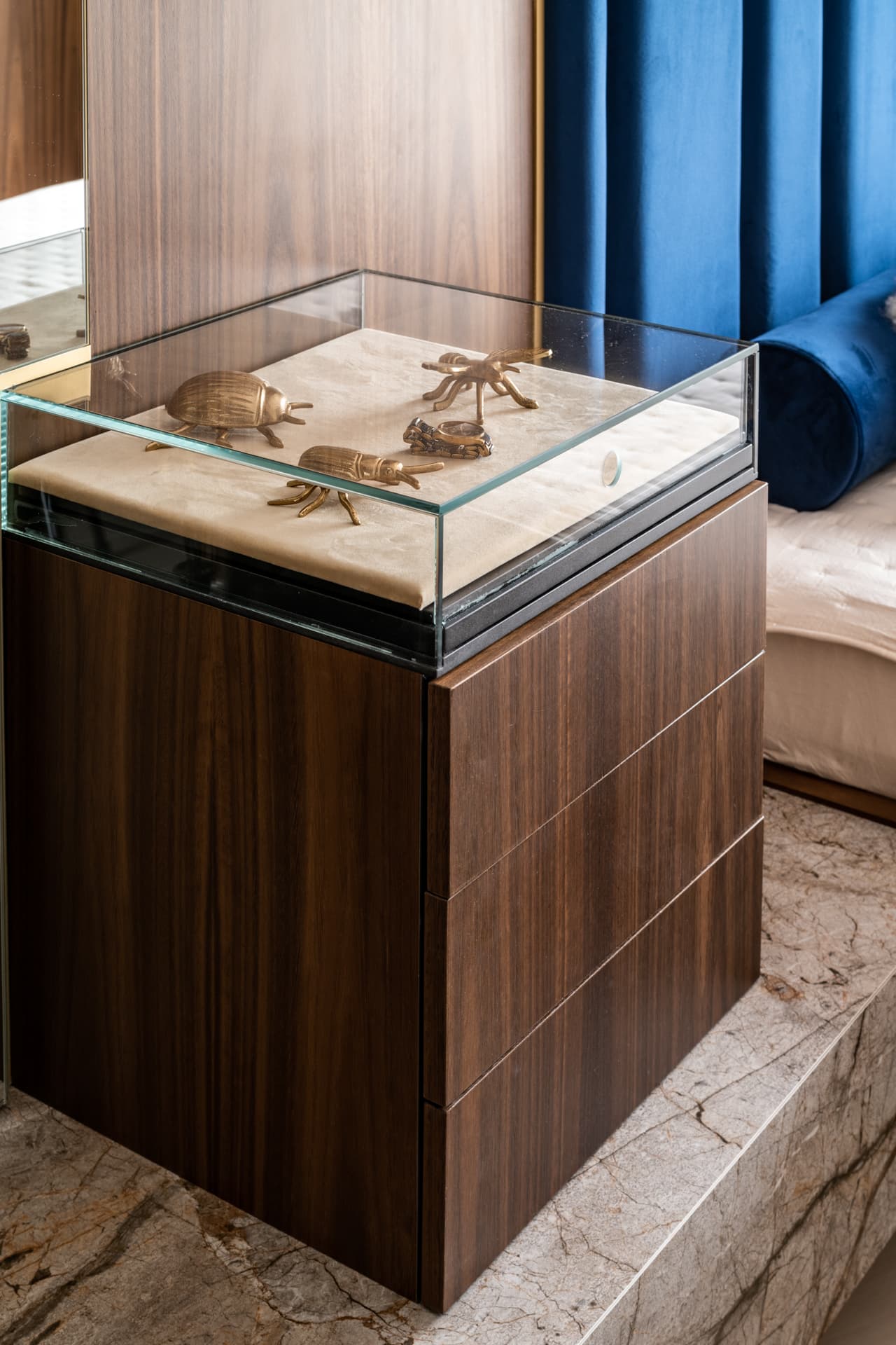 Glass-topped walnut display nightstand holding brass insect figurines on a stone plinth in the master bedroom of Argamán