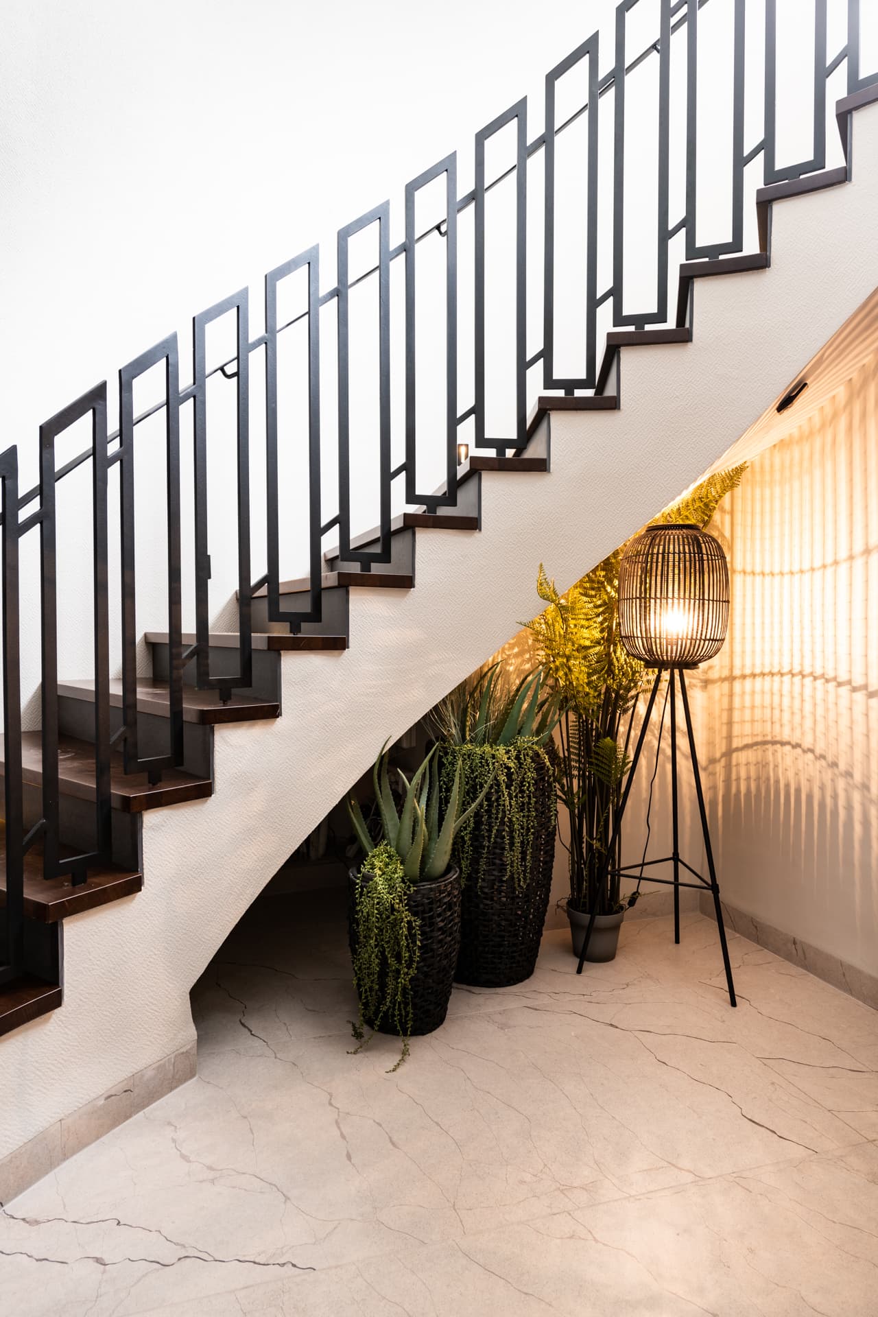 Staircase with a geometric black steel balustrade above potted plants and a woven lantern floor lamp in the entrance of Argamán