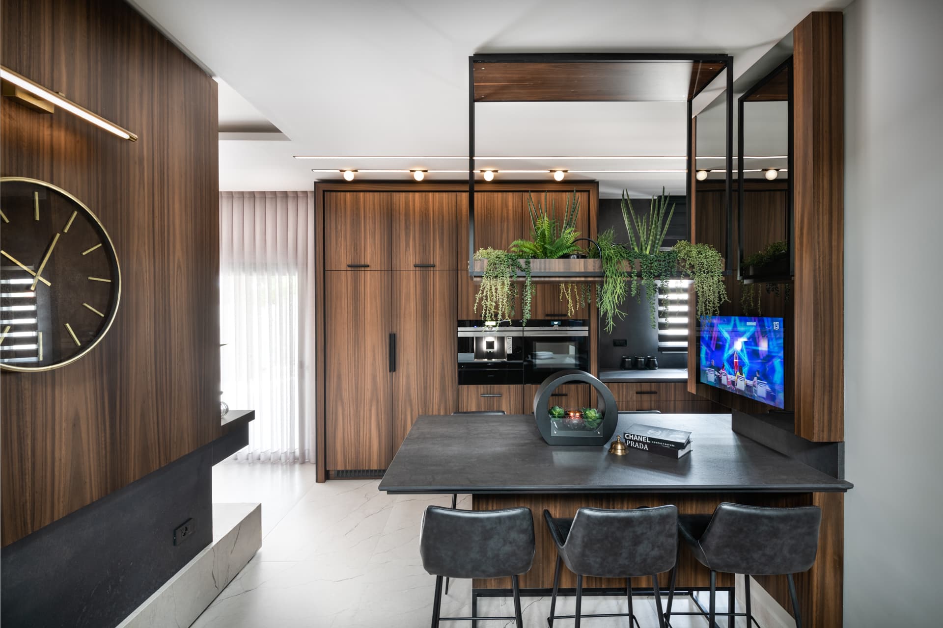 Kitchen island with a dark stone top and leather stools under a suspended steel planter frame, beside a round brass wall clock, in Argamán