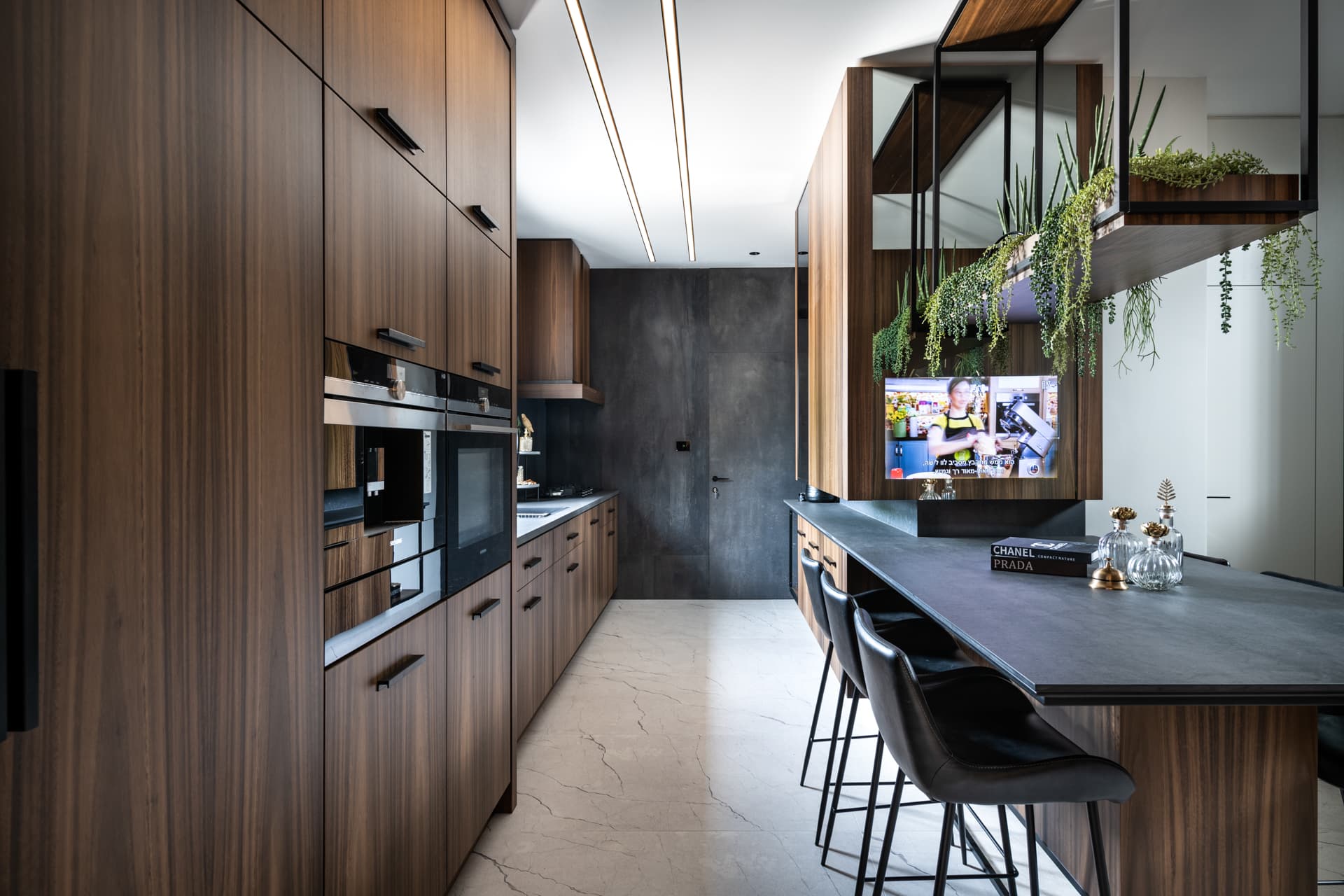 Galley view along walnut cabinetry toward a dark grey end wall, beside the island bar and hanging steel planter, in the kitchen of Argamán