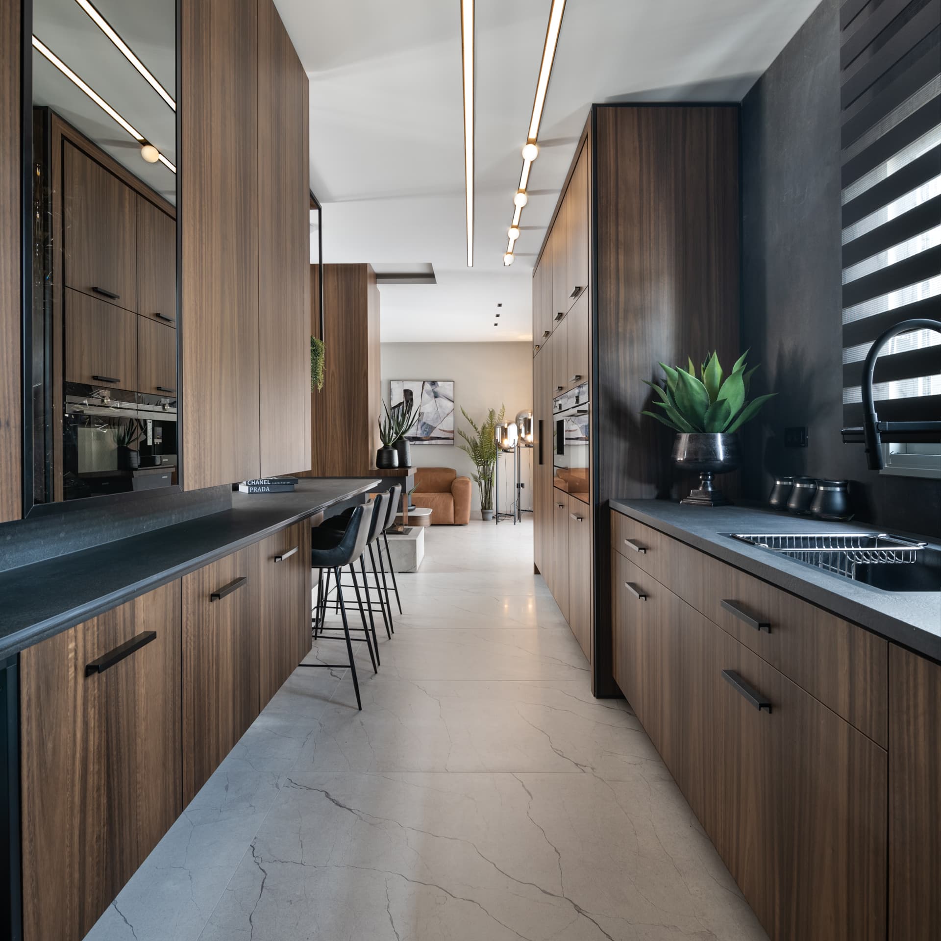 Long galley of walnut cabinets and dark stone worktops under linear track lights, opening to the living room, in the kitchen of Argamán