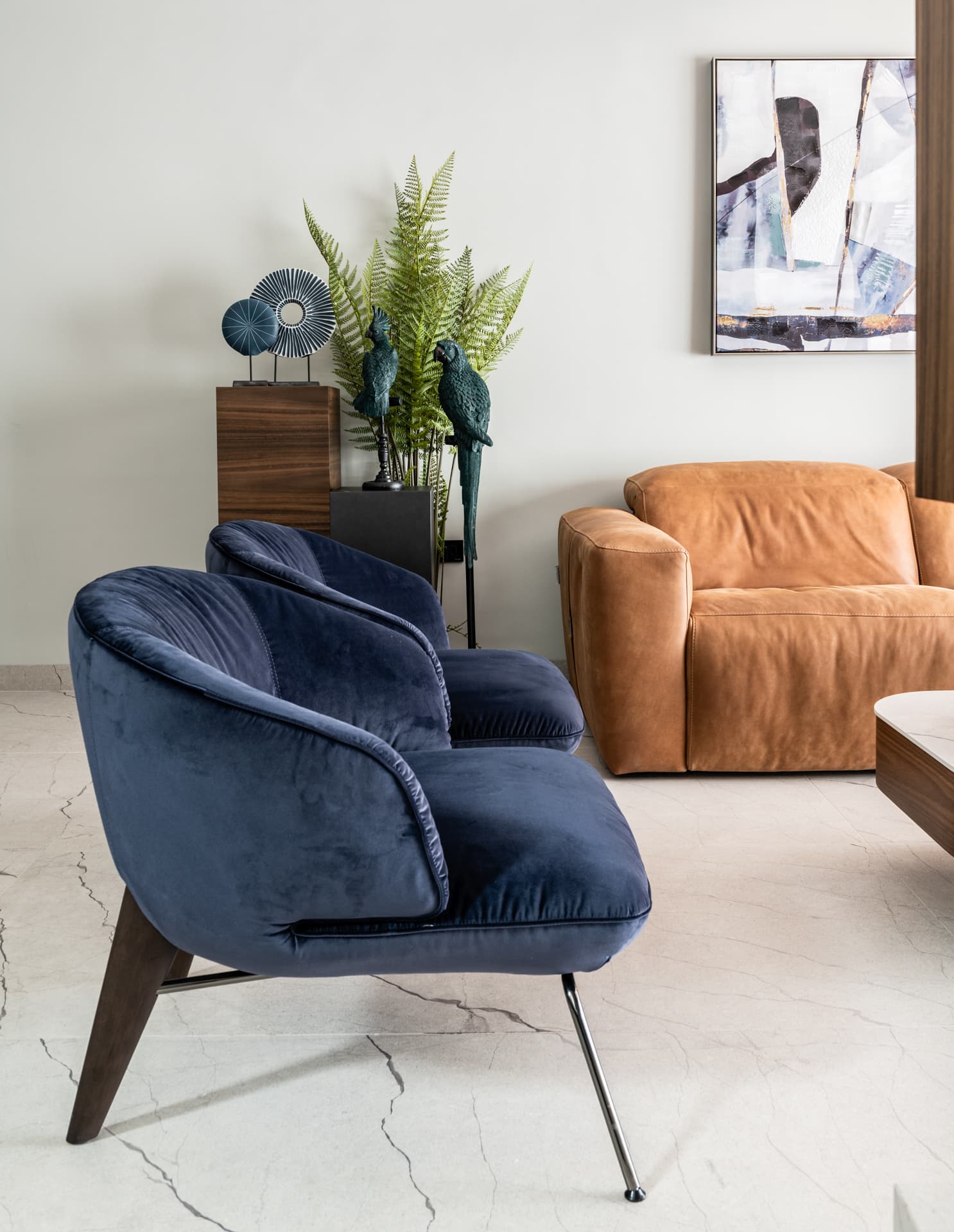 Pair of blue velvet lounge chairs beside a cognac leather sofa, with ferns and parrot figurines, in the living room of Argamán
