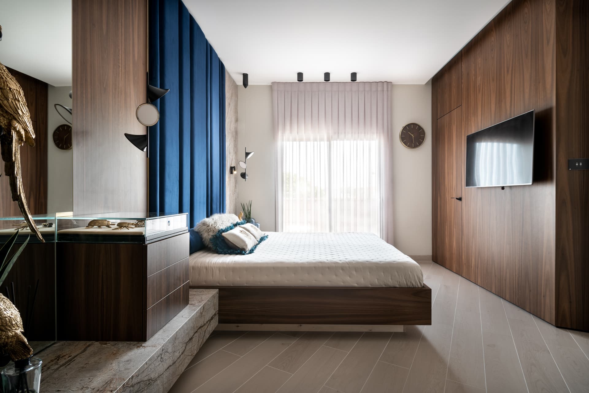 Daylight view of a floating walnut bed platform facing a walnut wardrobe wall with a mounted screen in the master bedroom of Argamán