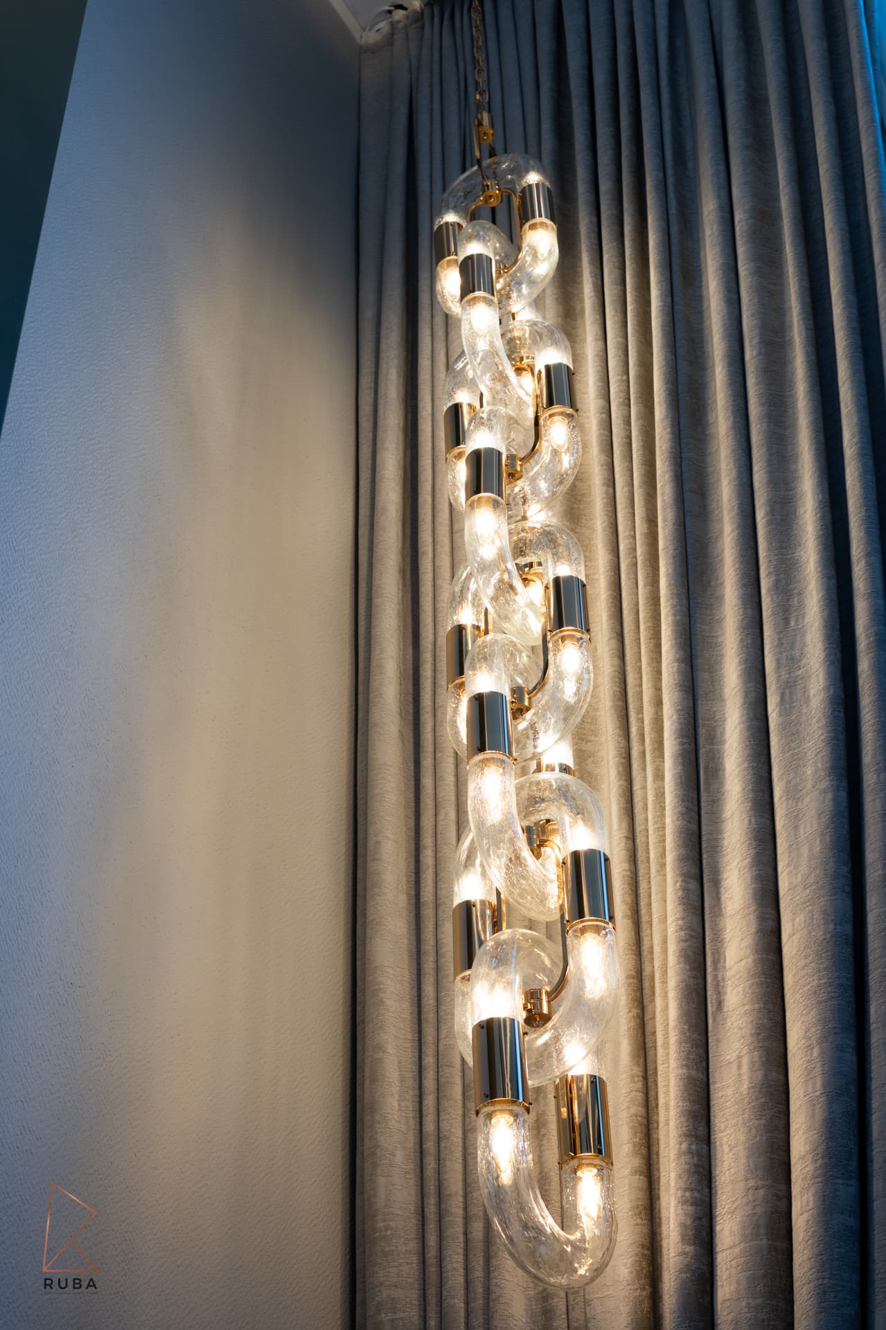 Glass chain-link pendant light with brass collars hanging against a grey curtain in the living room of Bird of Paradise Flower
