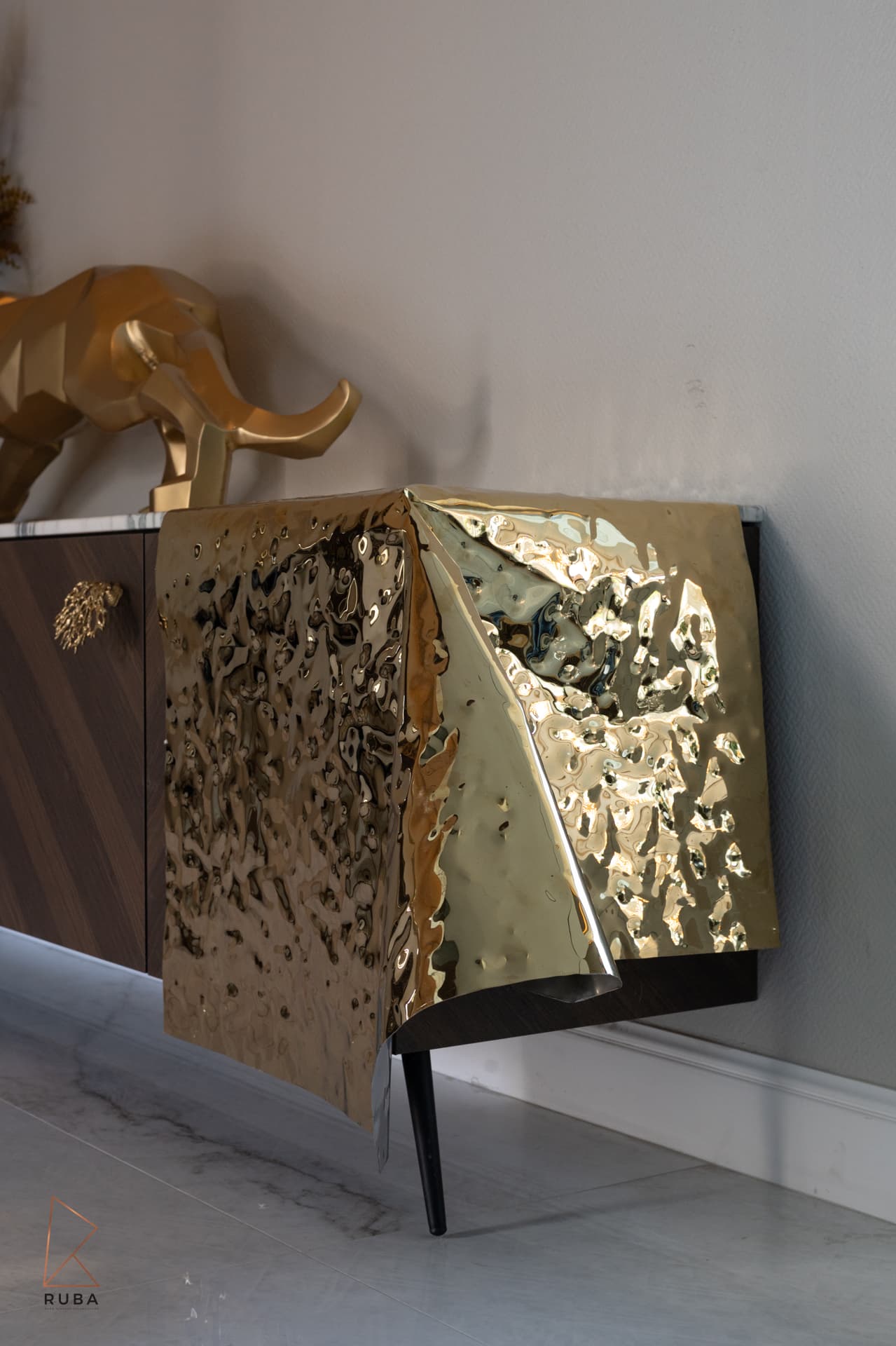 Hammered brass sheet draped over the end of a dark wood sideboard below a gold panther sculpture in Bird of Paradise Flower
