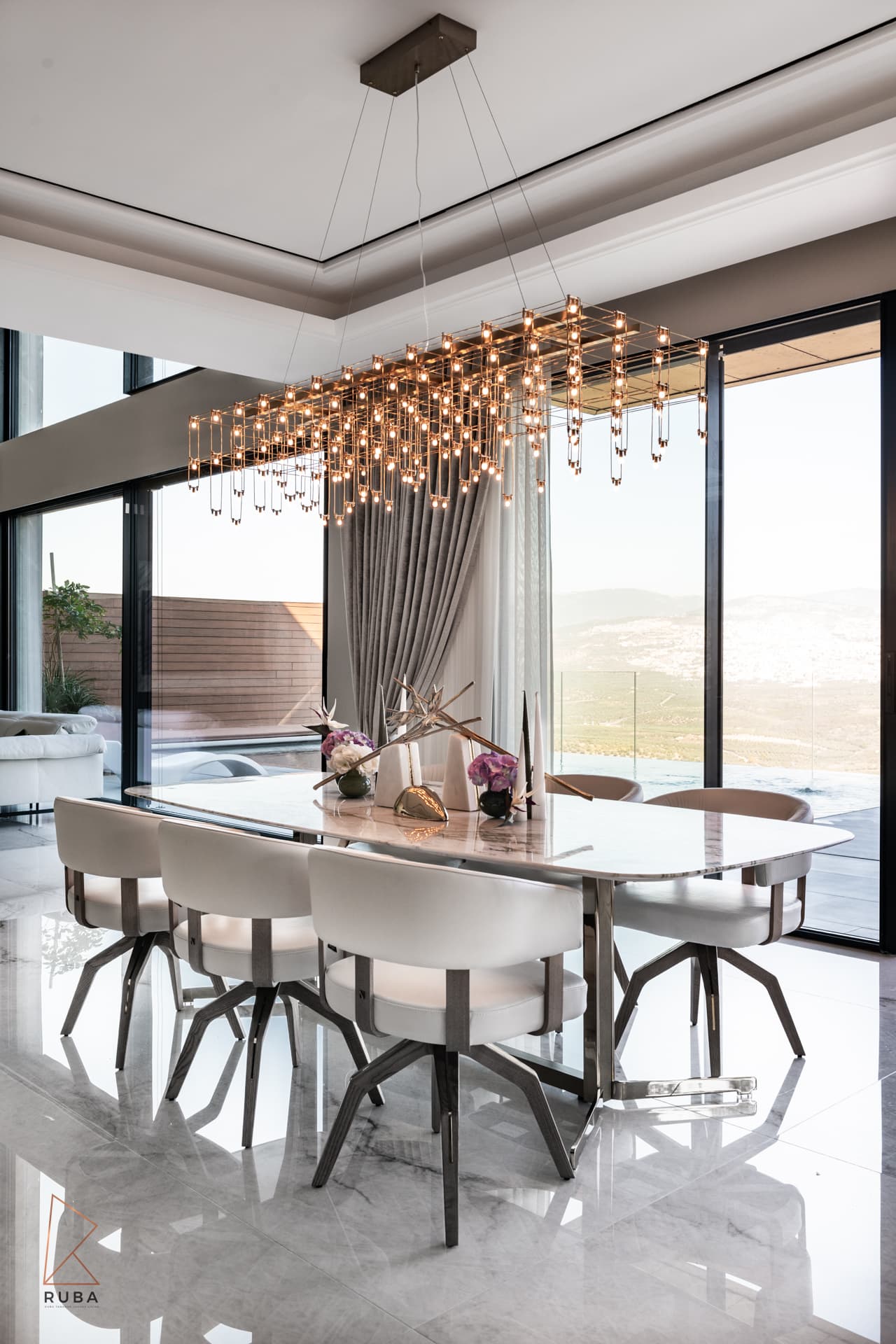 Dining room with a grid chandelier of hanging bulbs over a marble table and white curved chairs by tall windows in Bird of Paradise Flower