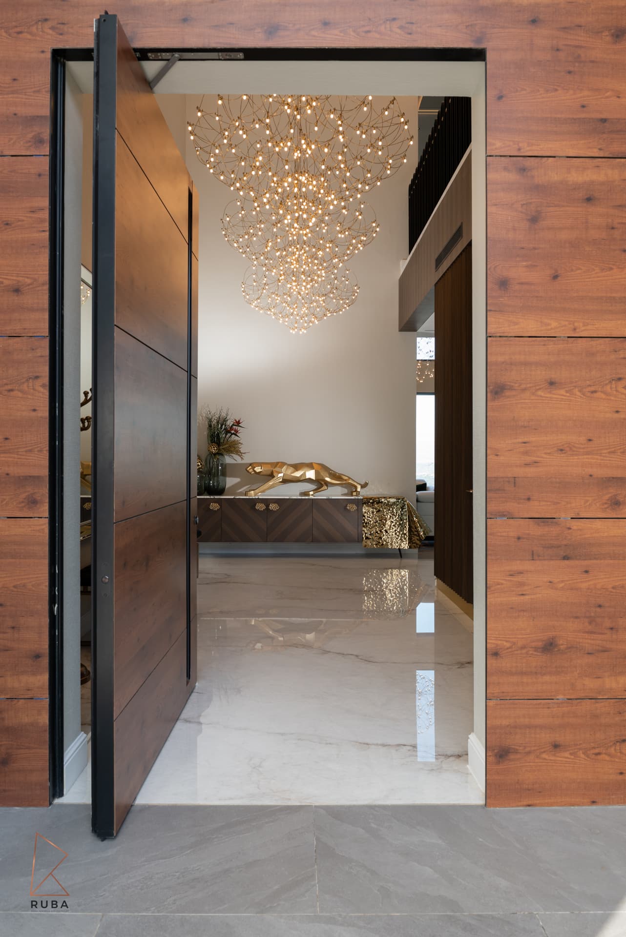 Open pivot front door in a wood-clad facade revealing a wire chandelier and panther sideboard in the entrance of Bird of Paradise Flower
