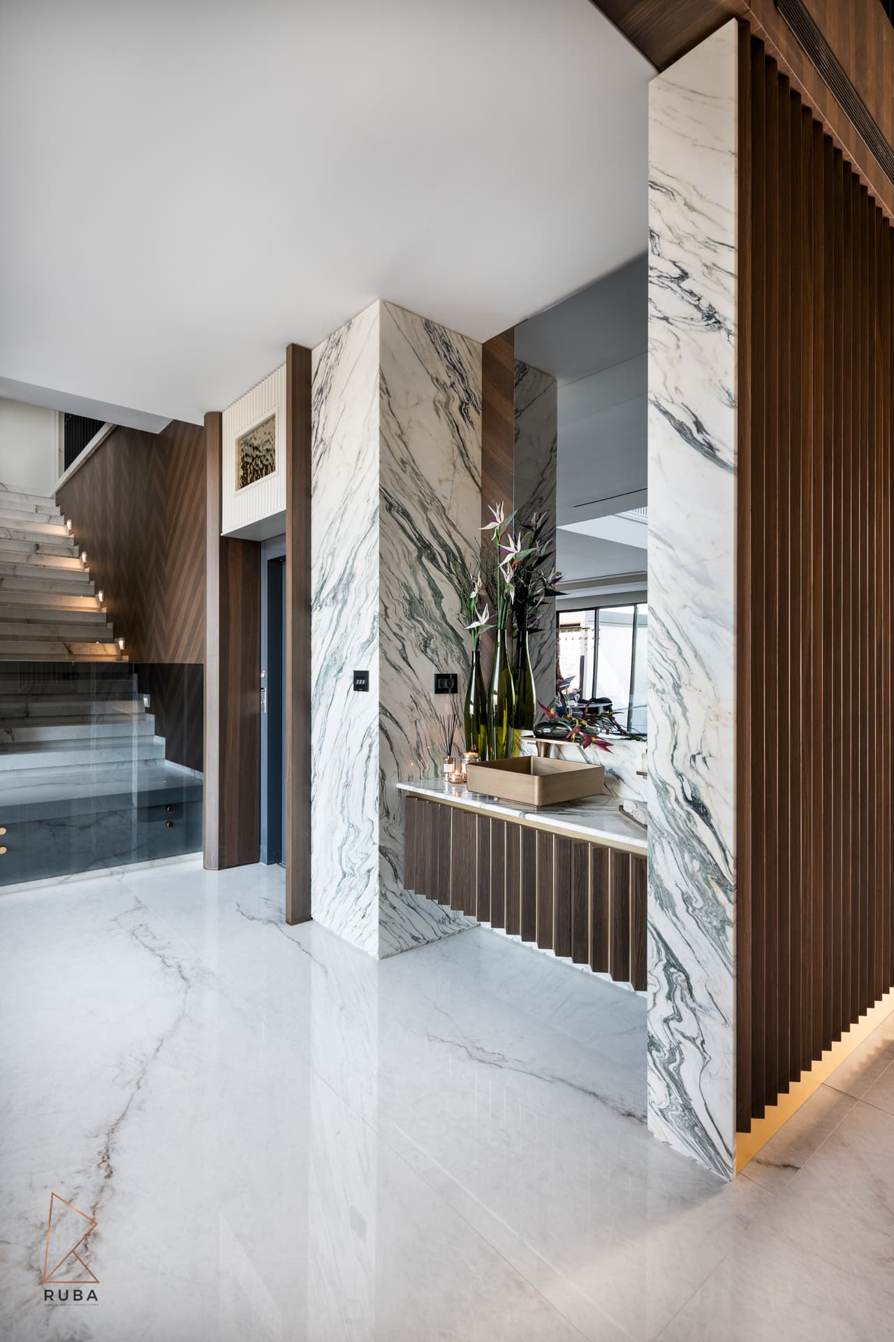 Veined marble-clad columns and a fluted wood console beside the glass-lined staircase in the entrance of Bird of Paradise Flower