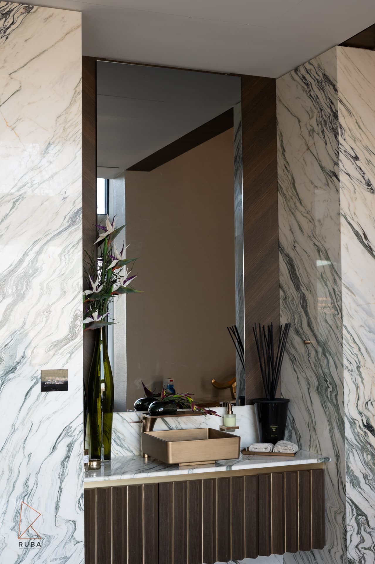 Veined marble walls framing a mirrored niche with a brushed brass basin on a fluted wood vanity in the entrance of Bird of Paradise Flower