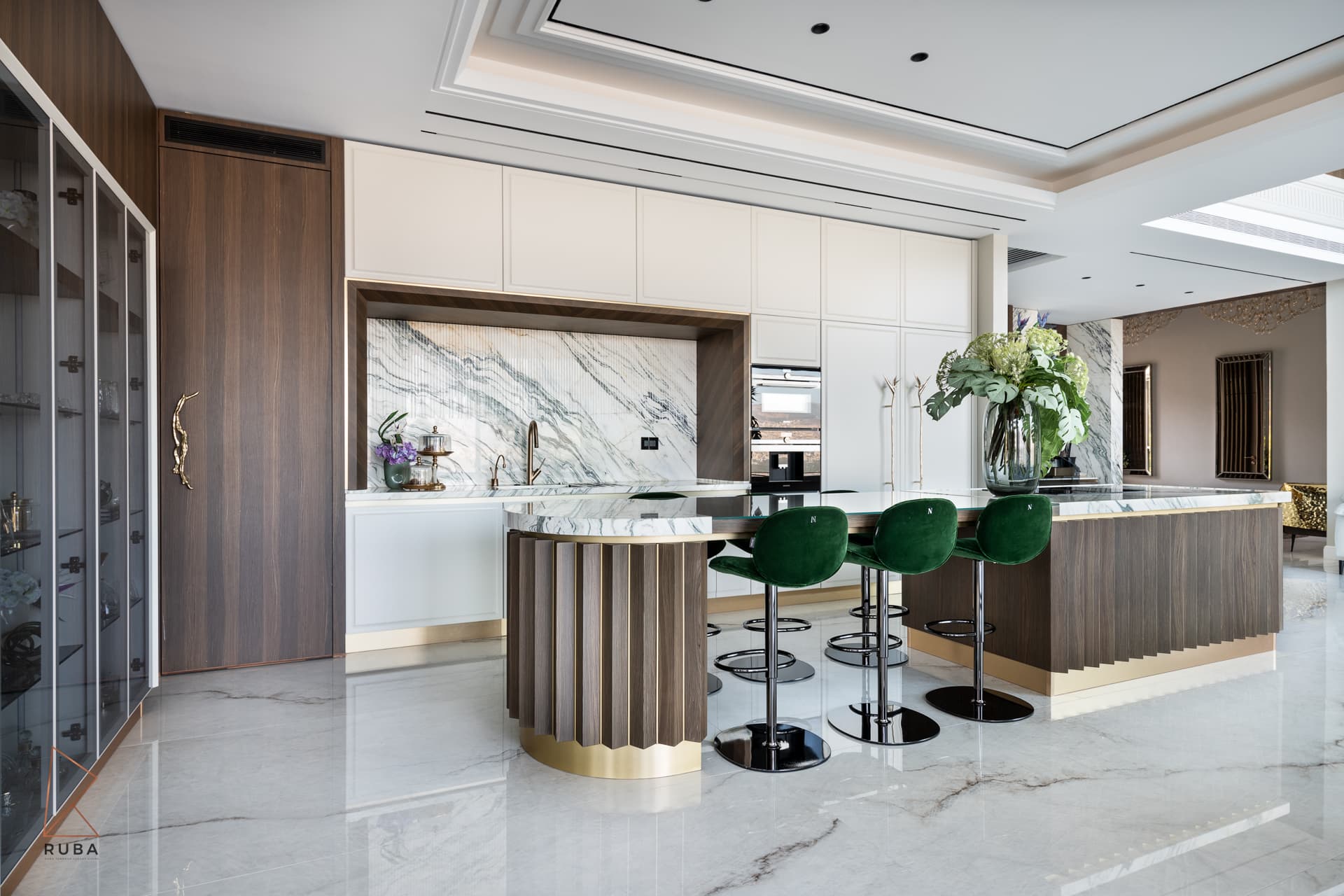 Kitchen island with a curved fluted wood end, marble top and three green velvet stools beside a glass vitrine in Bird of Paradise Flower