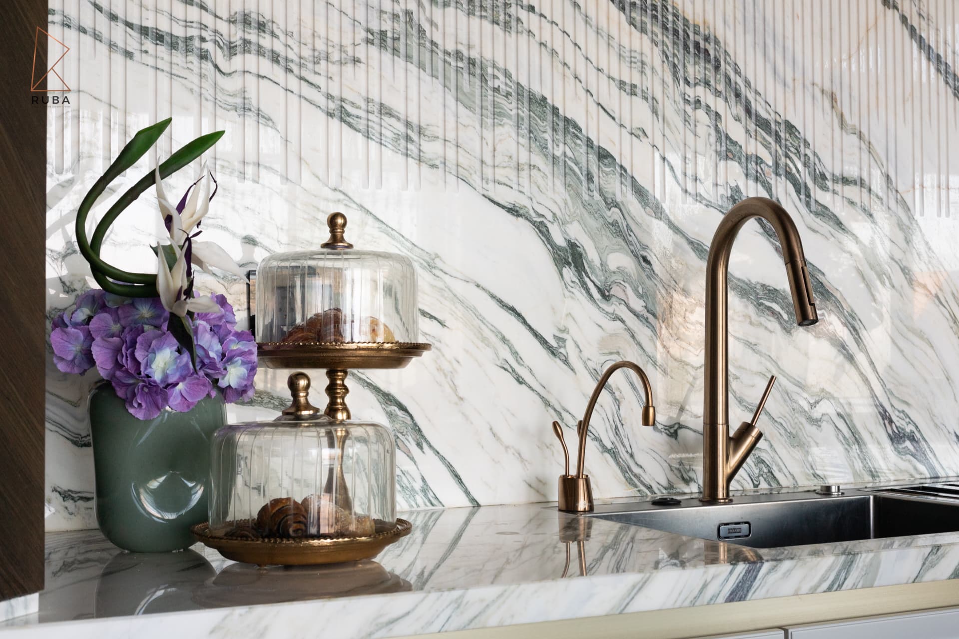 Bronze faucets and glass-domed brass cake stands against the fluted veined marble backsplash in the kitchen of Bird of Paradise Flower