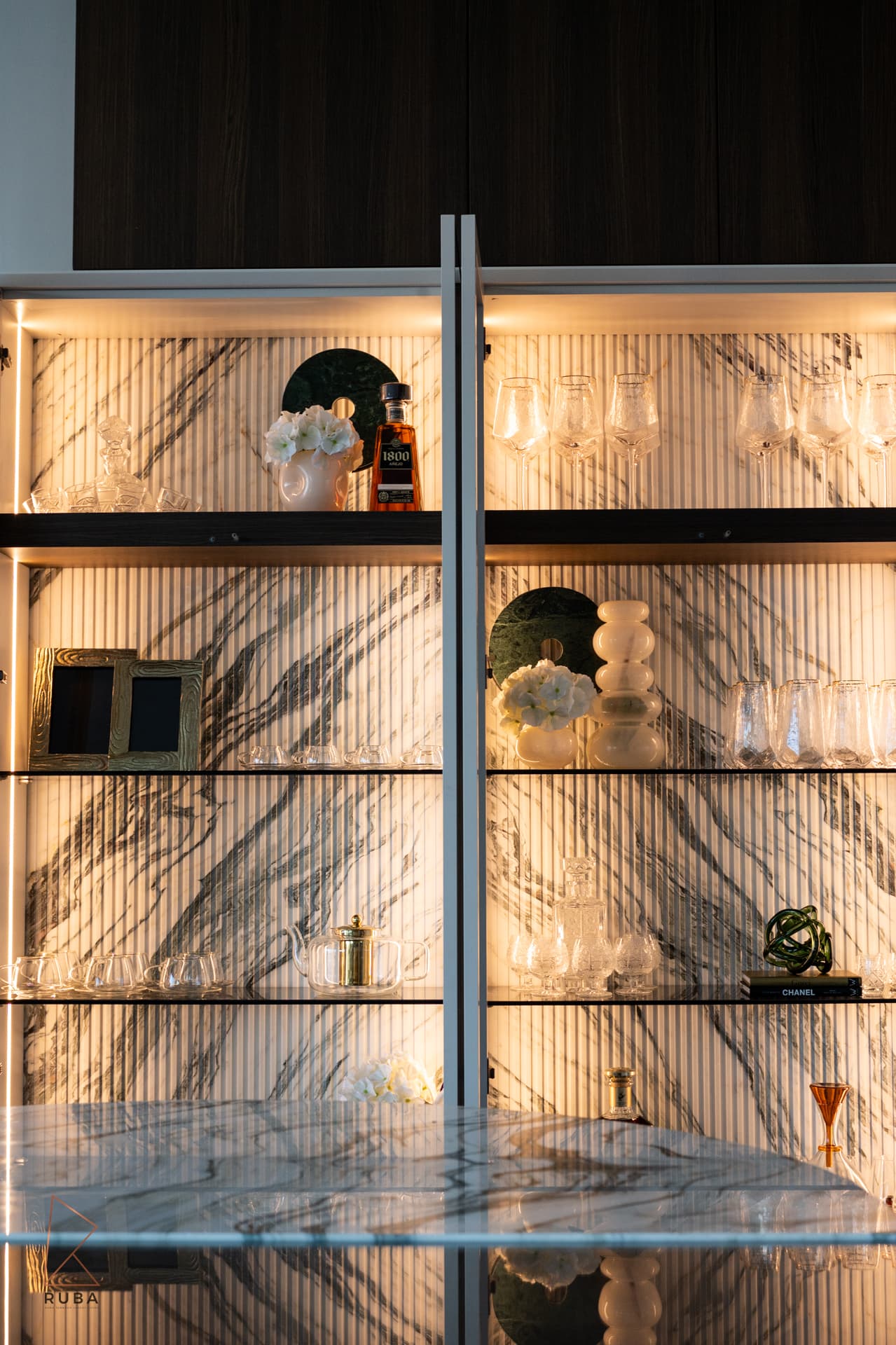 Backlit glass display cabinet with fluted veined marble backing and glassware on the shelves in the kitchen of Bird of Paradise Flower
