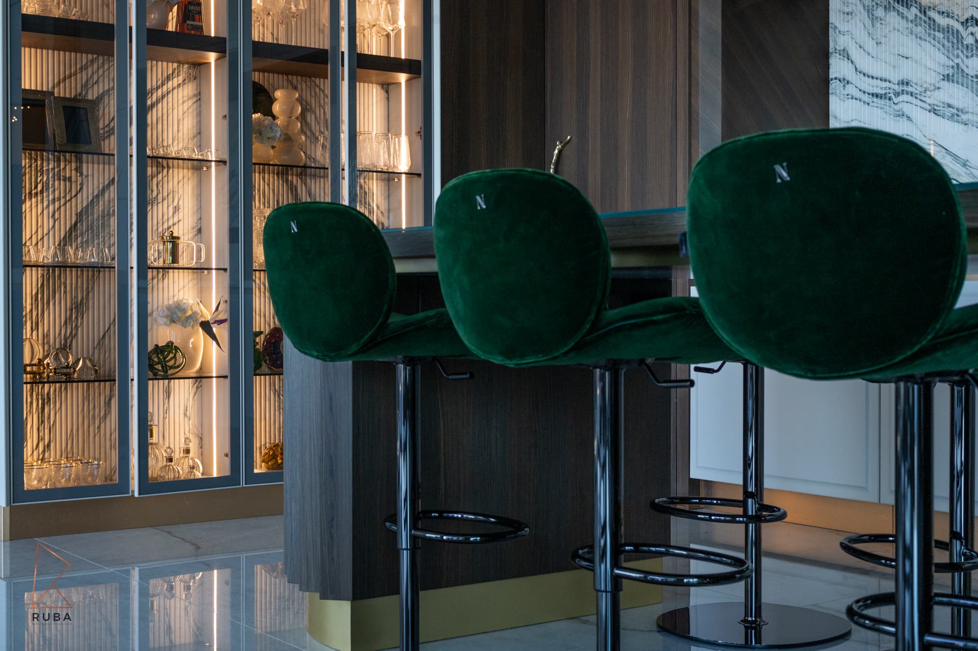 Row of green velvet bar stools at the dark wood island, with the lit display cabinet behind, in the kitchen of Bird of Paradise Flower