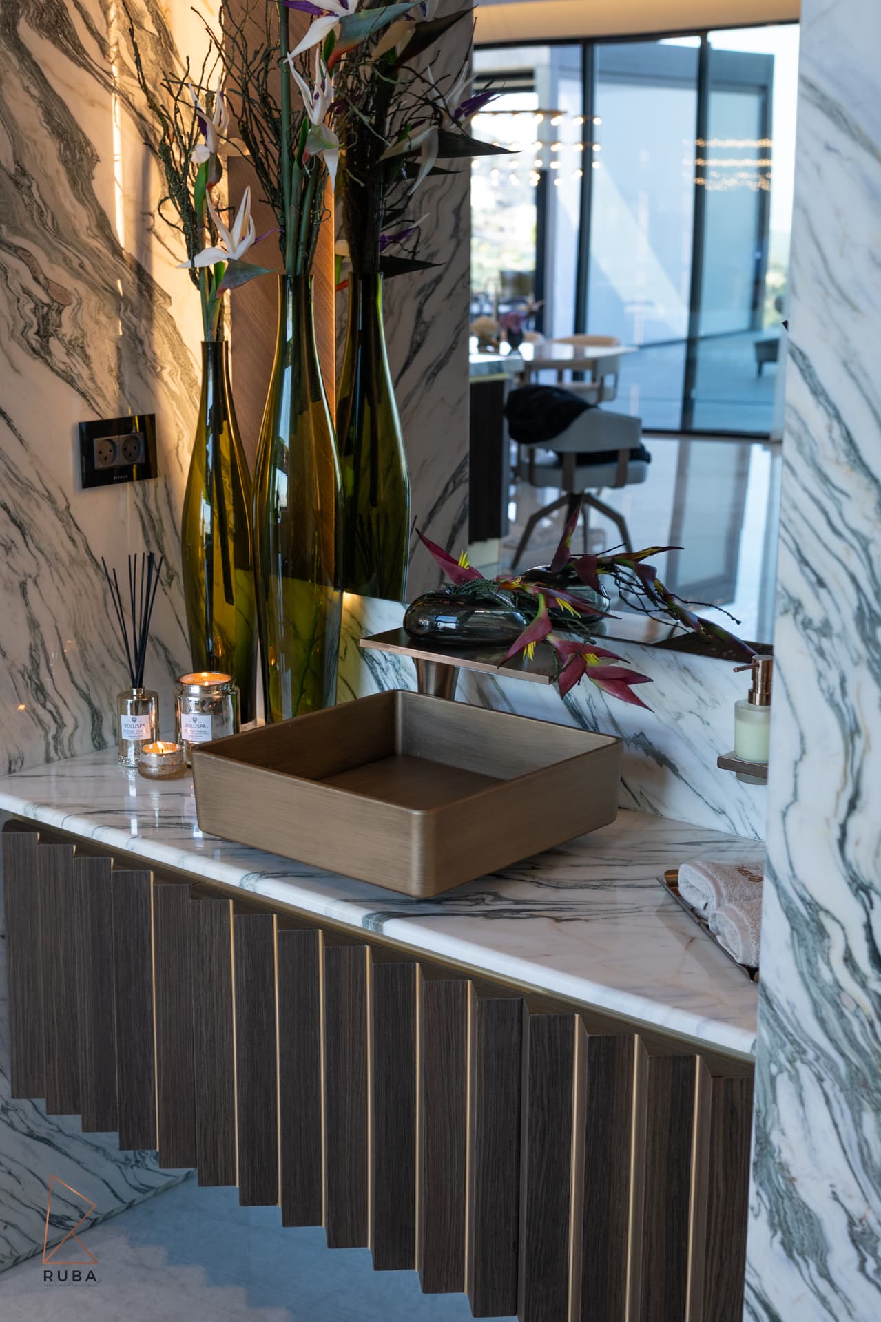 Side view of the brass basin, green glass vases and diffusers on the marble vanity in the powder room of Bird of Paradise Flower
