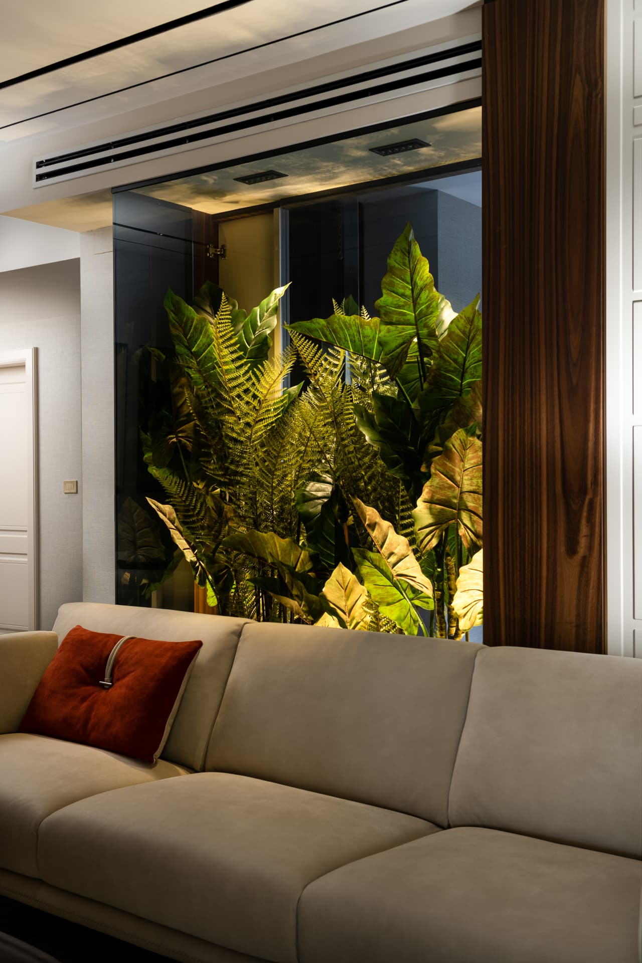 Glass planter niche filled with ferns and broad leaves behind a cream sofa with a rust cushion in Born Together