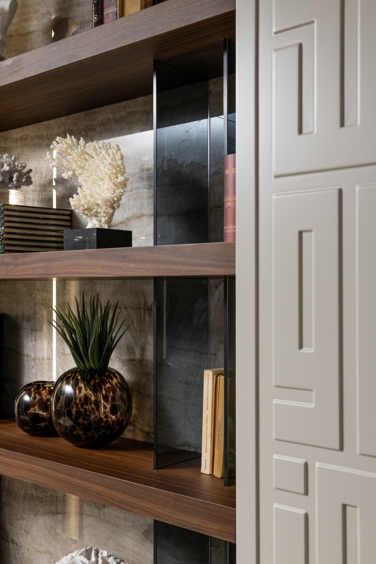 Walnut shelf with leopard-glass vases and white coral against veined stone beside a geometric relief panel in Born Together