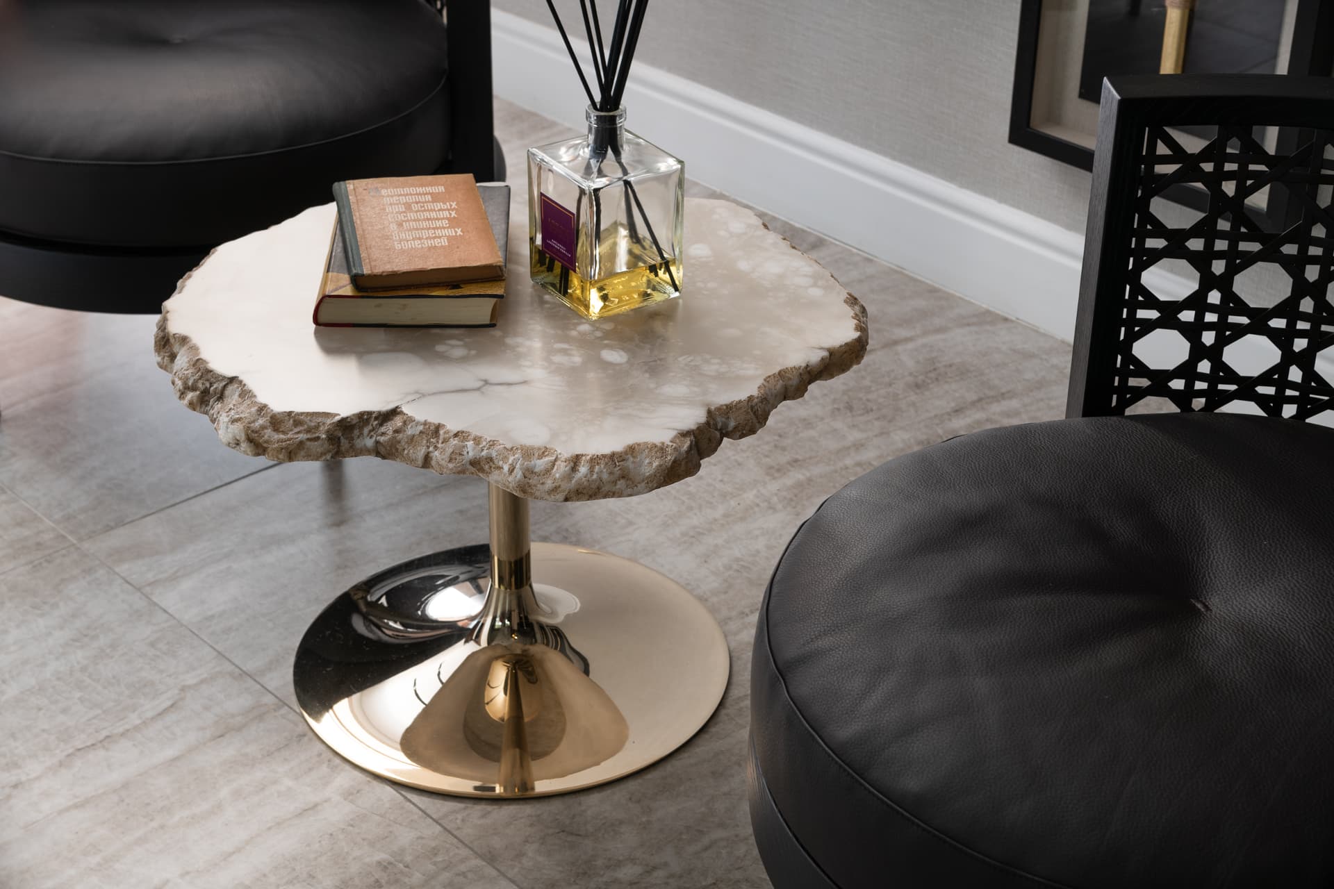 Raw-edge stone side table on a polished brass pedestal between black leather armchairs in Born Together