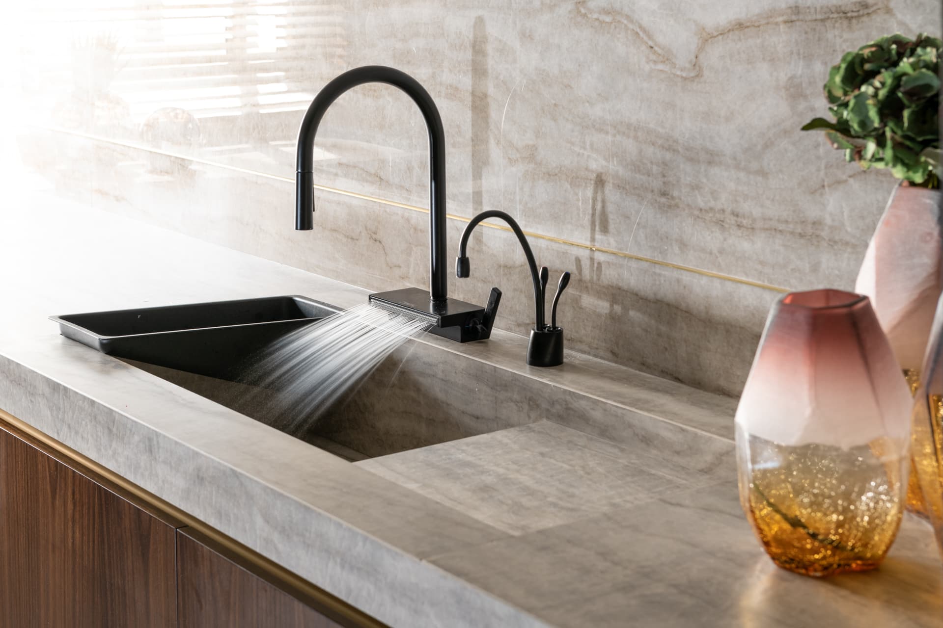 Black pull-out faucet over a black sink set into a sloping veined stone counter with a brass backsplash line in the kitchen of Born Together