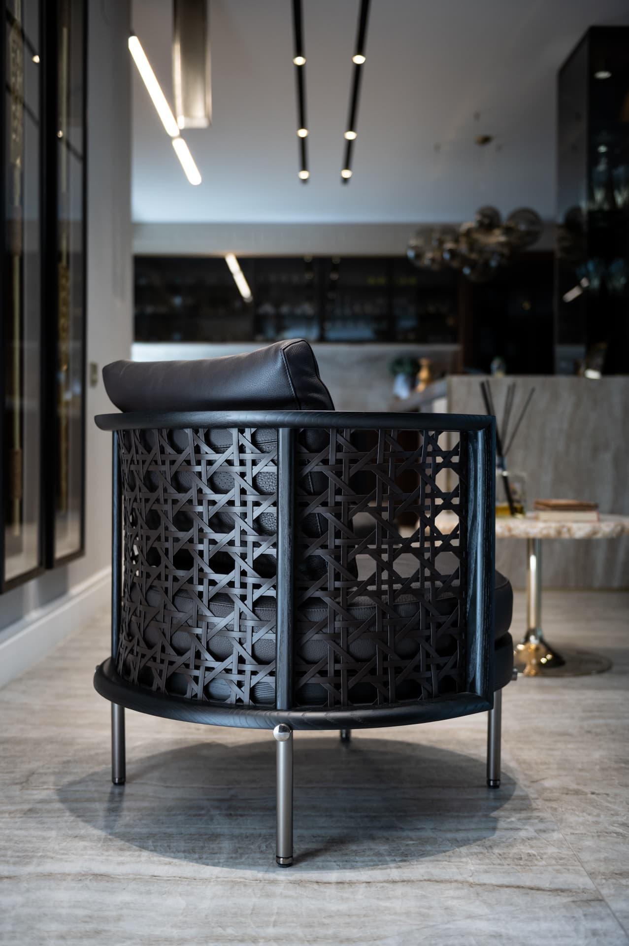Back of a black lattice armchair with leather cushions, seen toward the kitchen display wall in Born Together