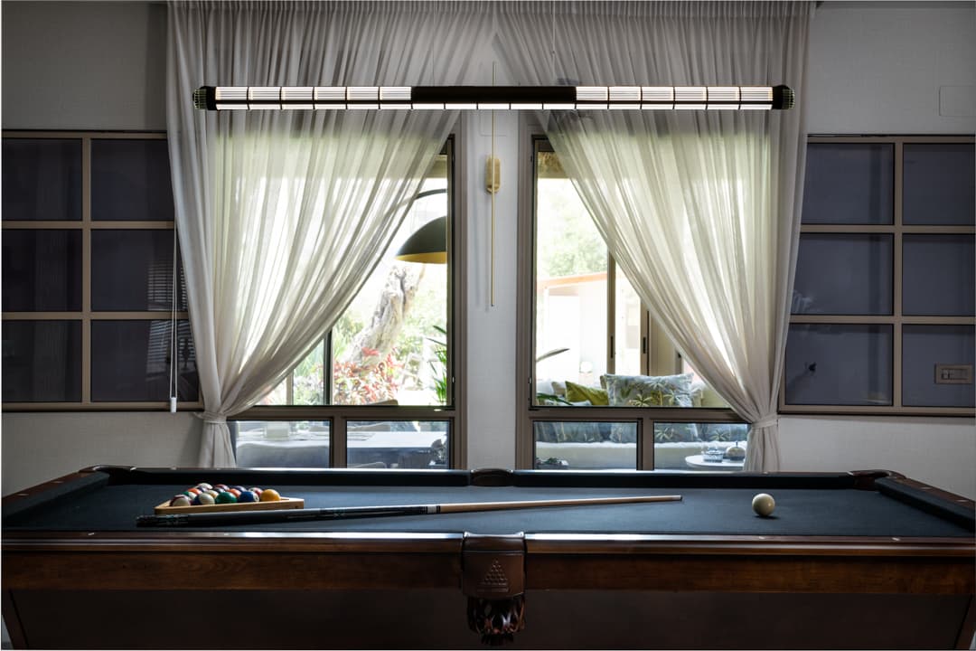 Game room with a billiard table beneath a long linear pendant, framed by sheer curtains at twin windows in Born Together