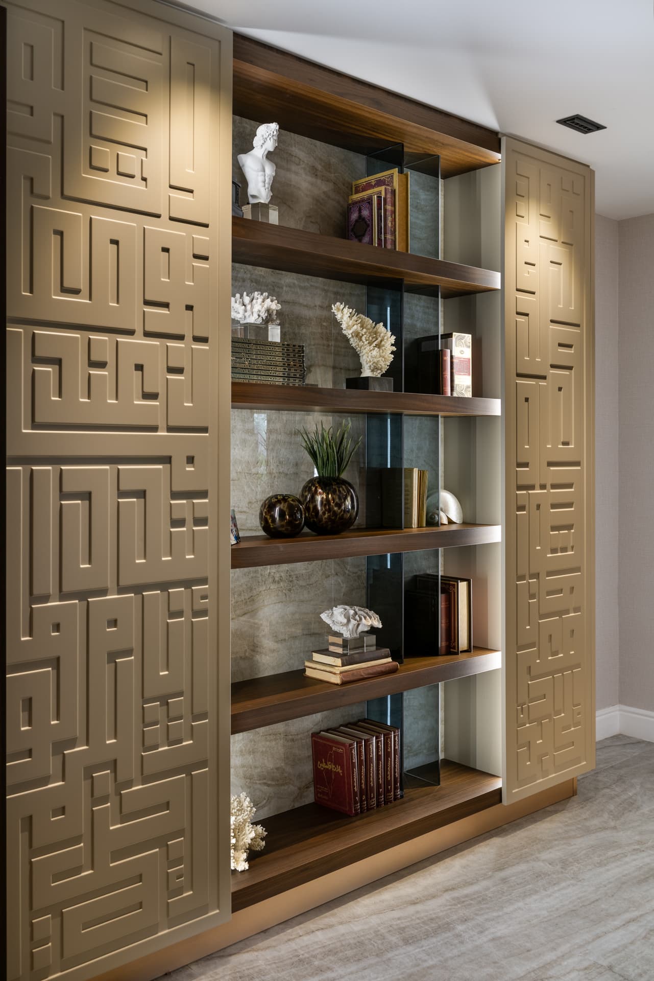 Walnut shelves with smoked glass fins and stone backing flanked by maze-like geometric relief panels in the game room of Born Together