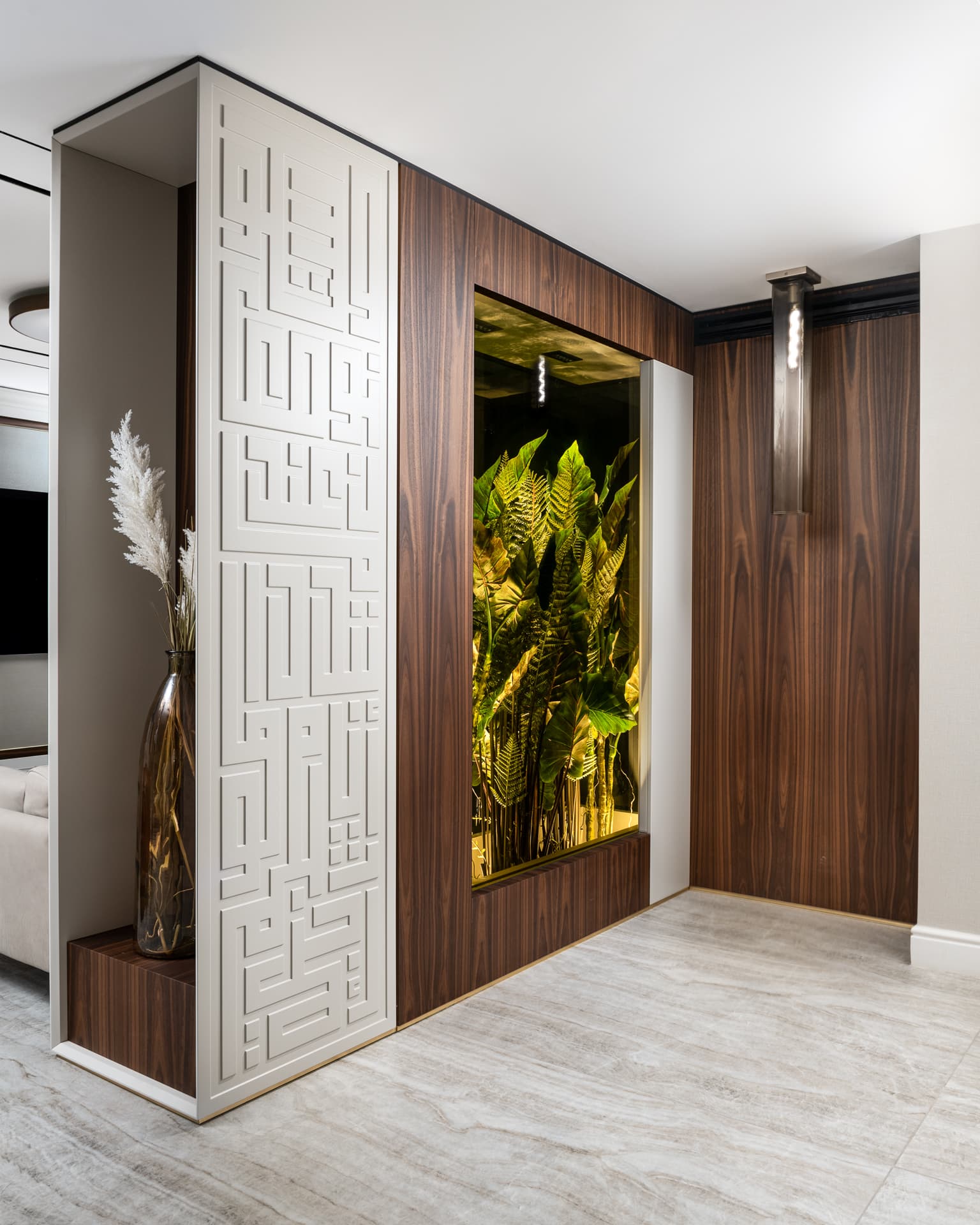 Hallway with a white geometric relief column, walnut-clad wall and a lit glass planter niche in Born Together