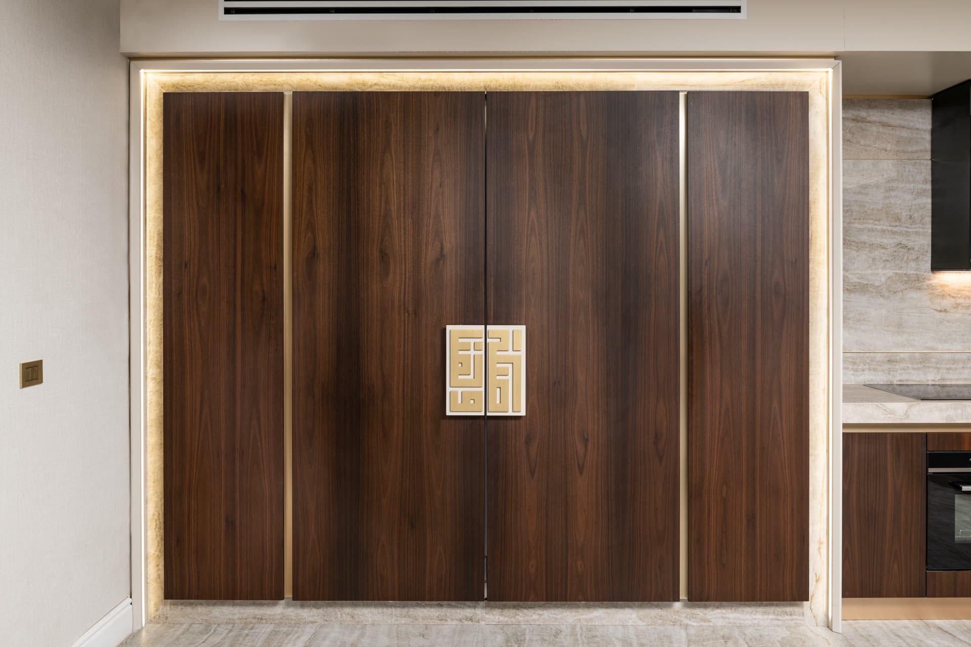 Hallway wall of walnut doors in a backlit stone frame with a brass geometric pull at the center in Born Together