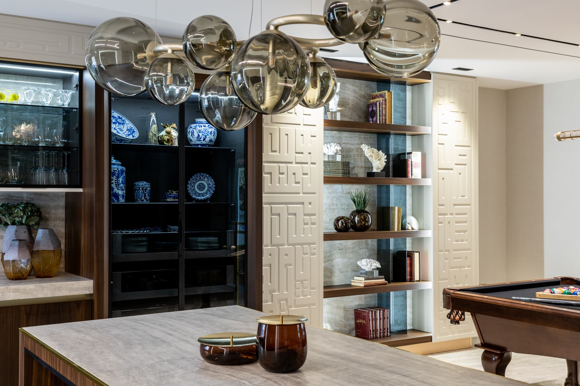 Kitchen island in veined stone under a smoked-glass globe chandelier, facing lit display shelving in Born Together