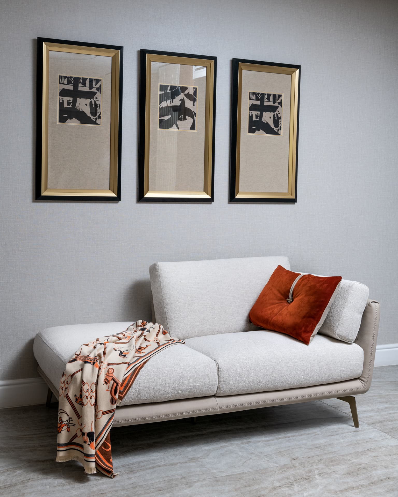Cream chaise sofa with a rust cushion beneath three gold-framed prints in the living room of Born Together
