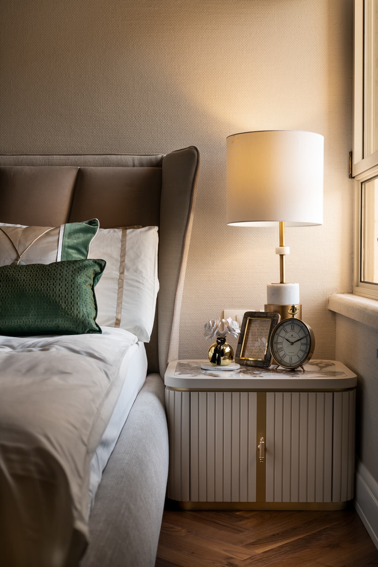 Side view of the tufted headboard beside a fluted nightstand with a table lamp and a clock by the window in the bedroom of Cognac