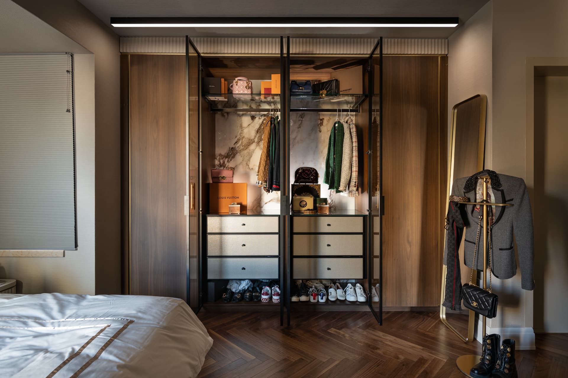 Open glass-door wardrobe with a veined stone back panel set into walnut cabinetry above a herringbone floor in the bedroom of Cognac
