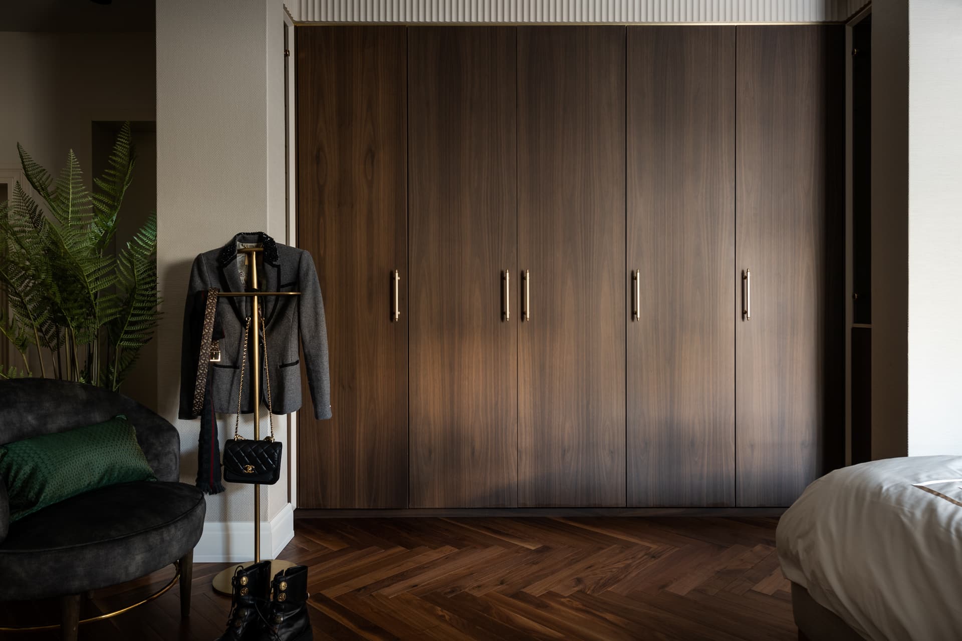 Wall of walnut wardrobes with brass pulls above a herringbone wood floor, with a valet stand and armchair, in the bedroom of Cognac
