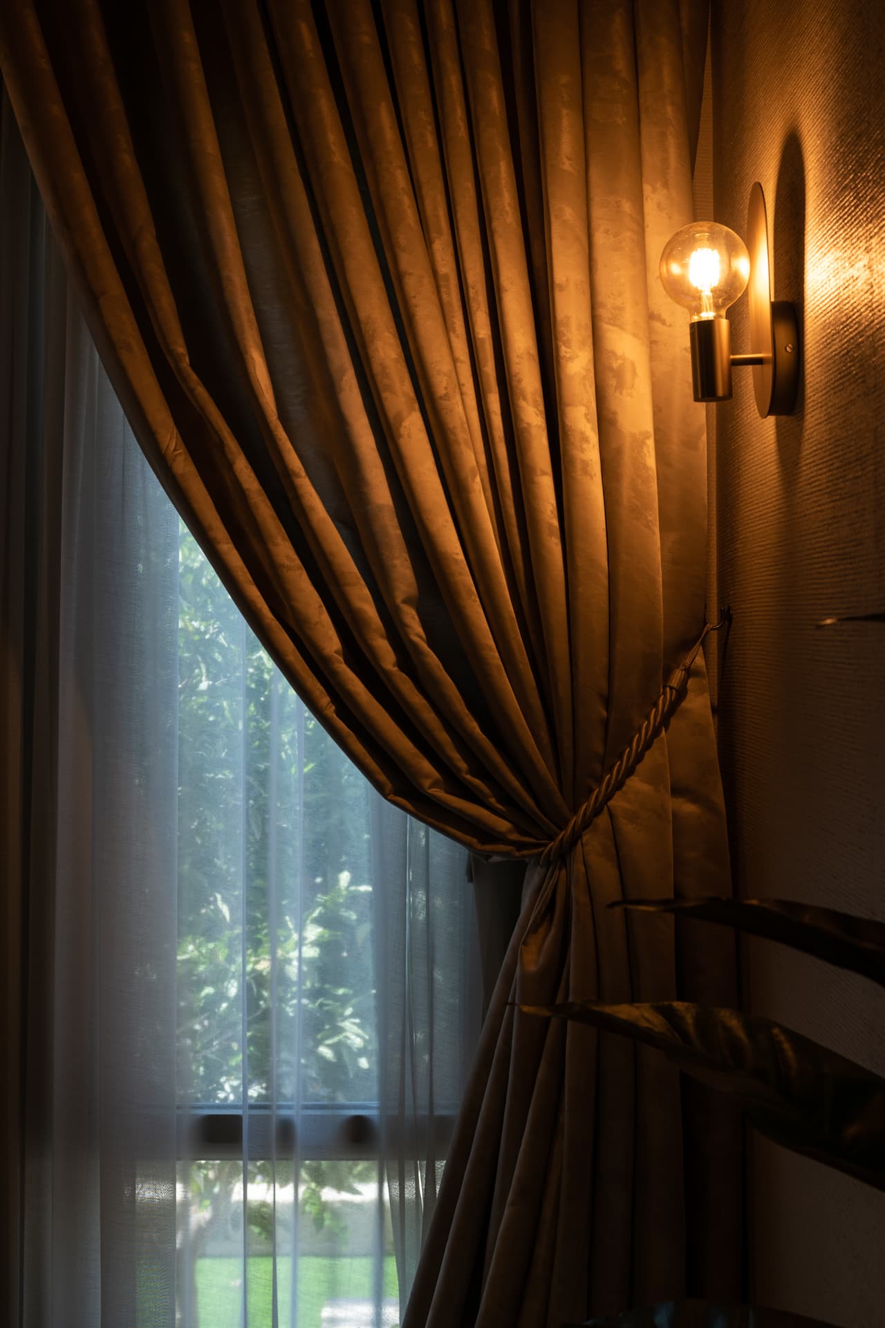 Exposed-bulb wall sconce beside a gathered curtain tied back with a rope, over a sheer panel, in the dining room of Cognac