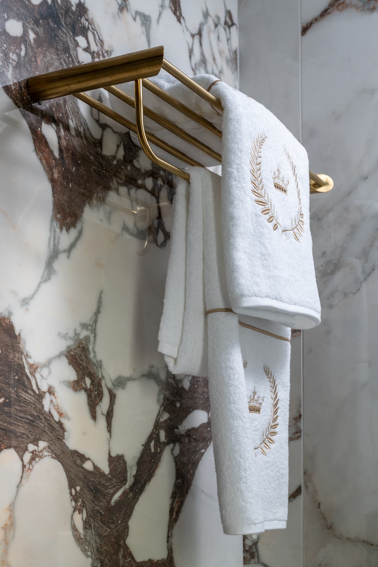 Brushed brass towel rack holding gold-embroidered white towels against dark-veined white stone in the master bathroom of Cognac