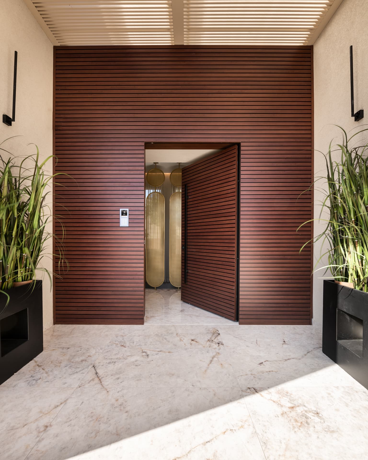 Horizontal wood-slat entrance wall with an open slatted door revealing brass oval screens inside, under a slatted canopy, at Cognac