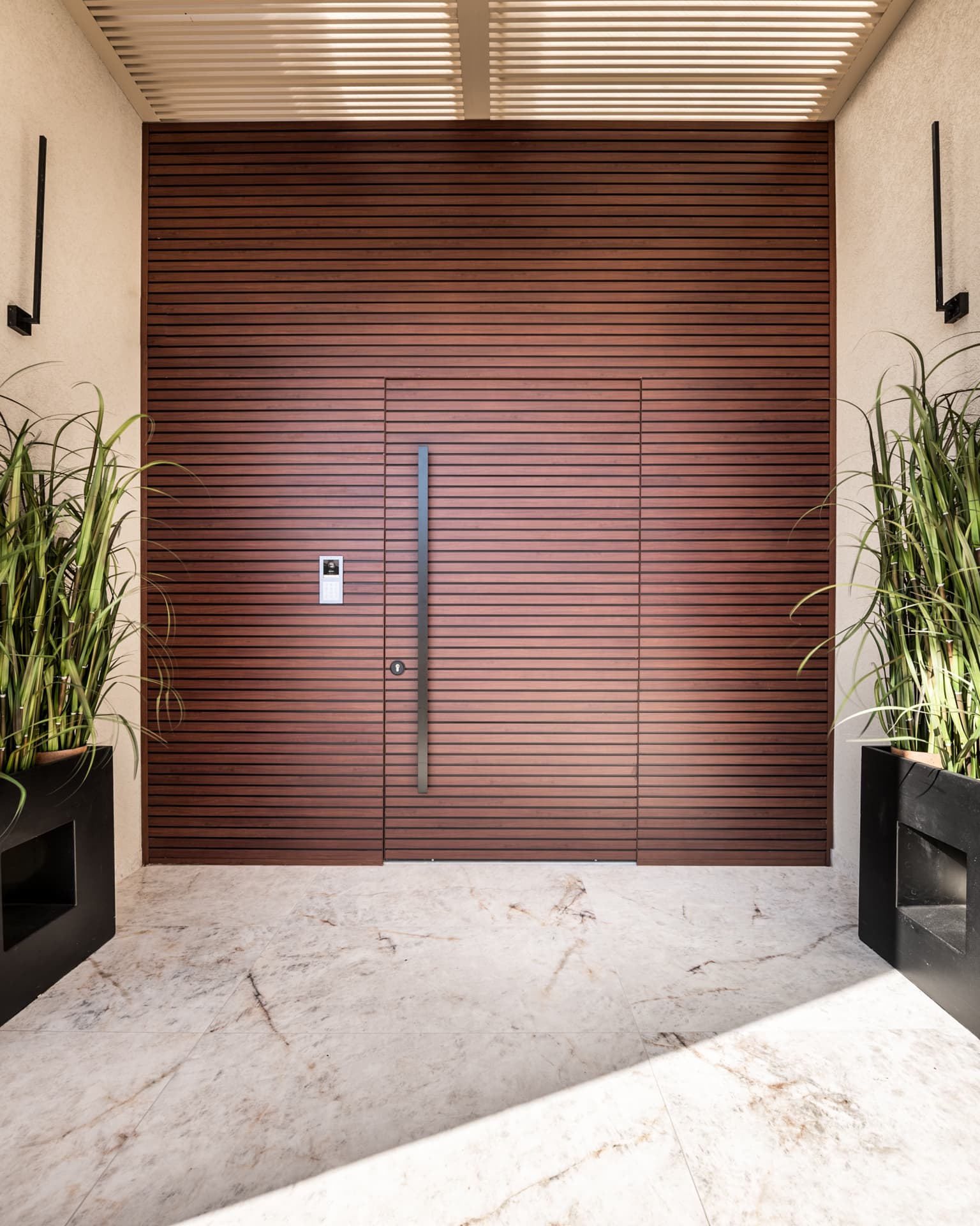 Closed front door with a long vertical pull, flush within a horizontal wood-slat wall and flanked by grass planters, at Cognac