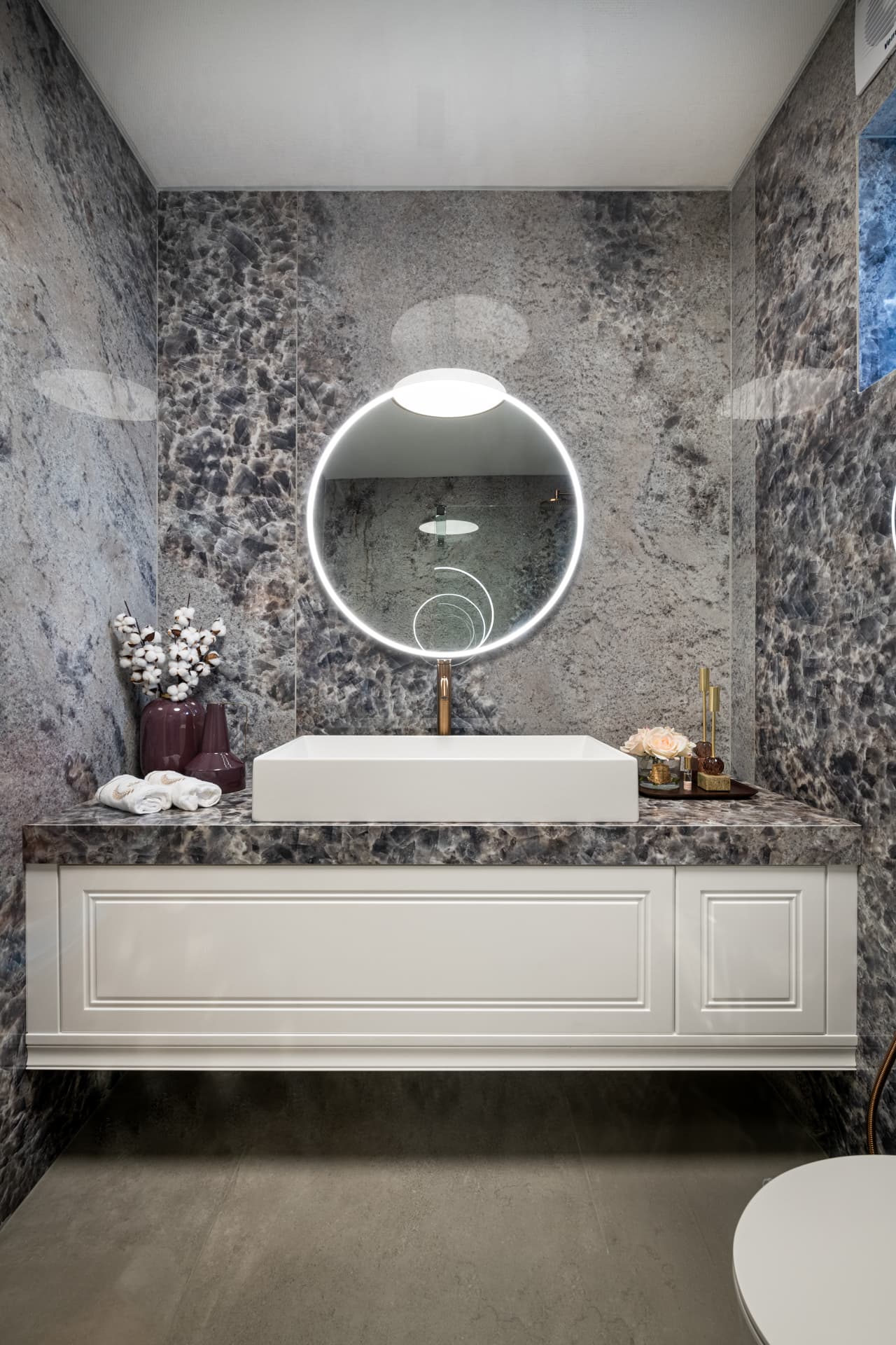 Floating white vanity with a vessel basin and a backlit round mirror against speckled blue-grey stone walls in the guest bathroom of Cognac