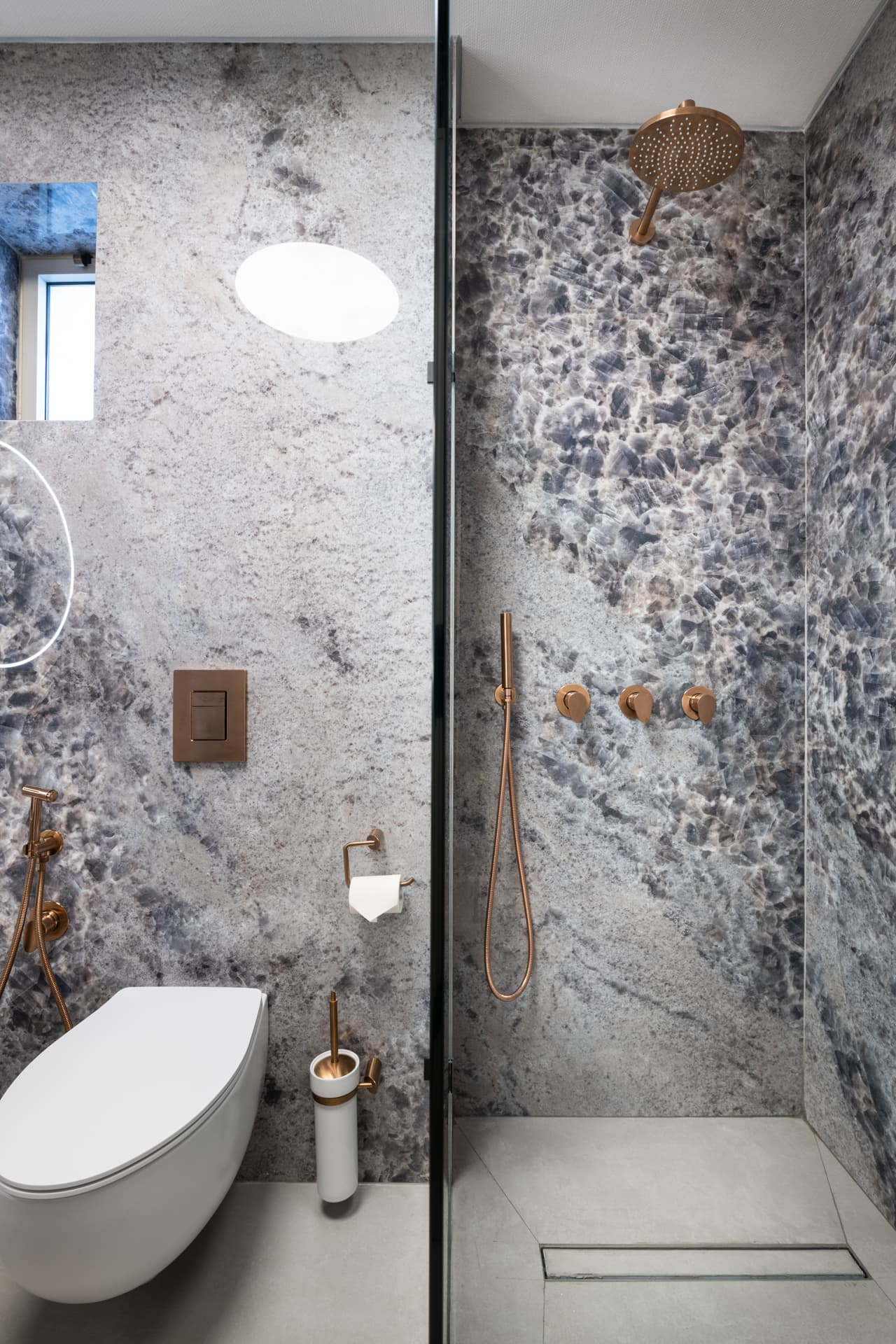 Walk-in shower with copper-toned fixtures behind glass beside a wall-hung toilet in blue-grey stone in the guest bathroom of Cognac