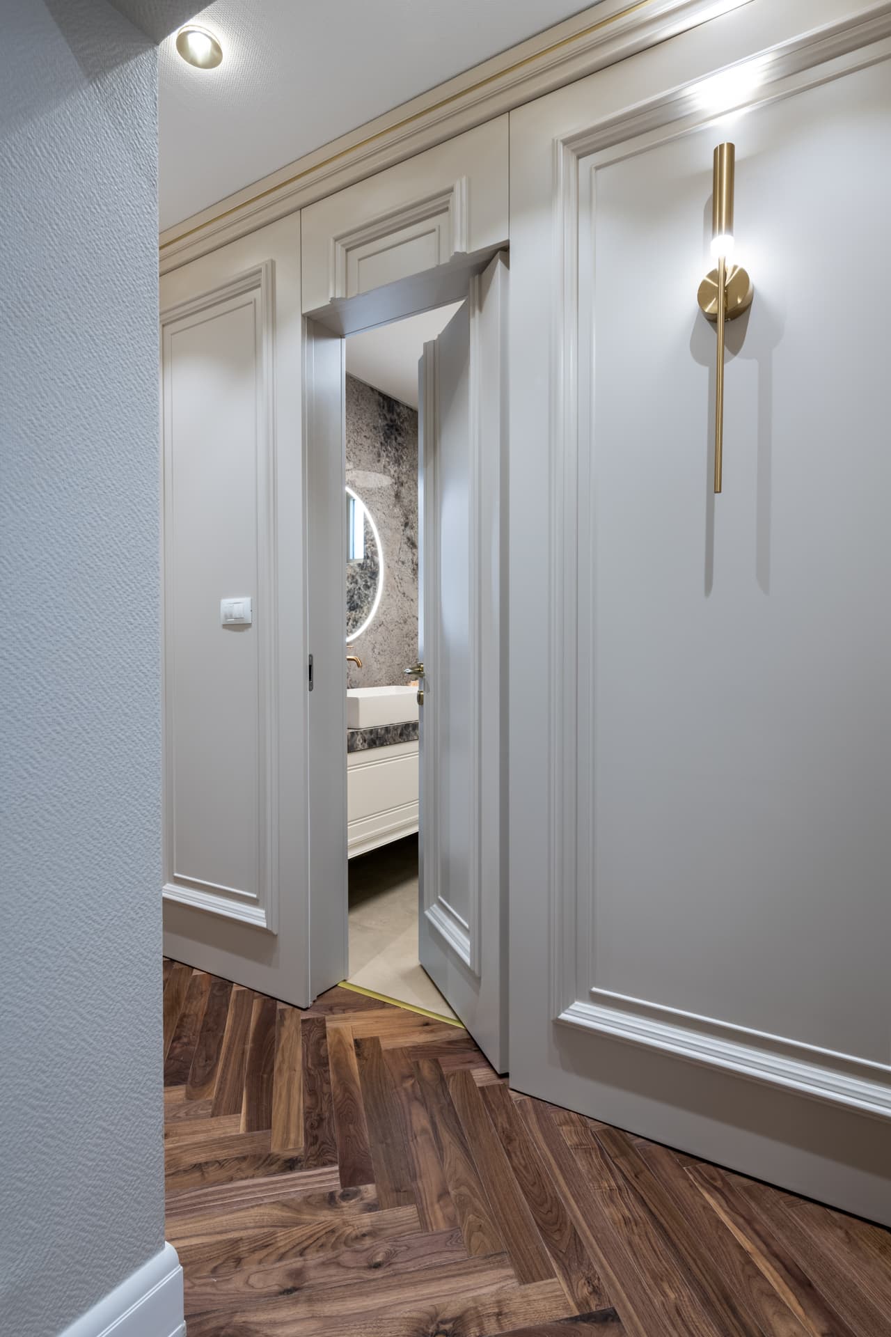 Paneled hallway with a brass stick wall sconce, herringbone walnut floor and a door opening to the guest bathroom in Cognac