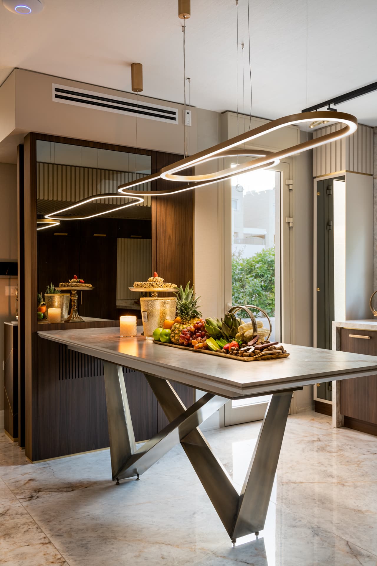 Veined stone kitchen table on angular bronze legs beneath a looped ring pendant, with a mirrored walnut wall behind, in Cognac