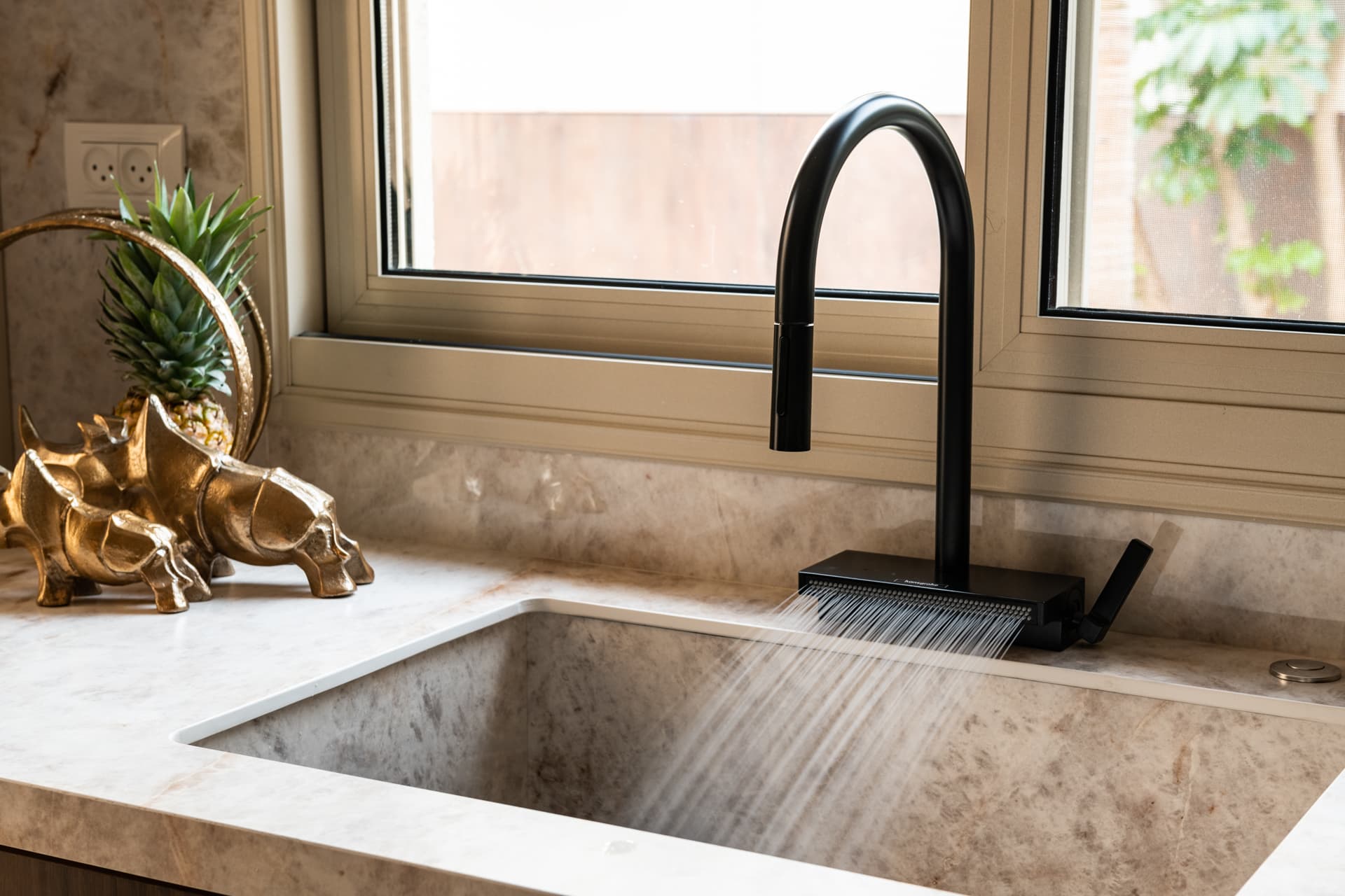 Black pull-out faucet over an integrated veined stone sink below a window, beside a brass animal sculpture, in the kitchen of Cognac