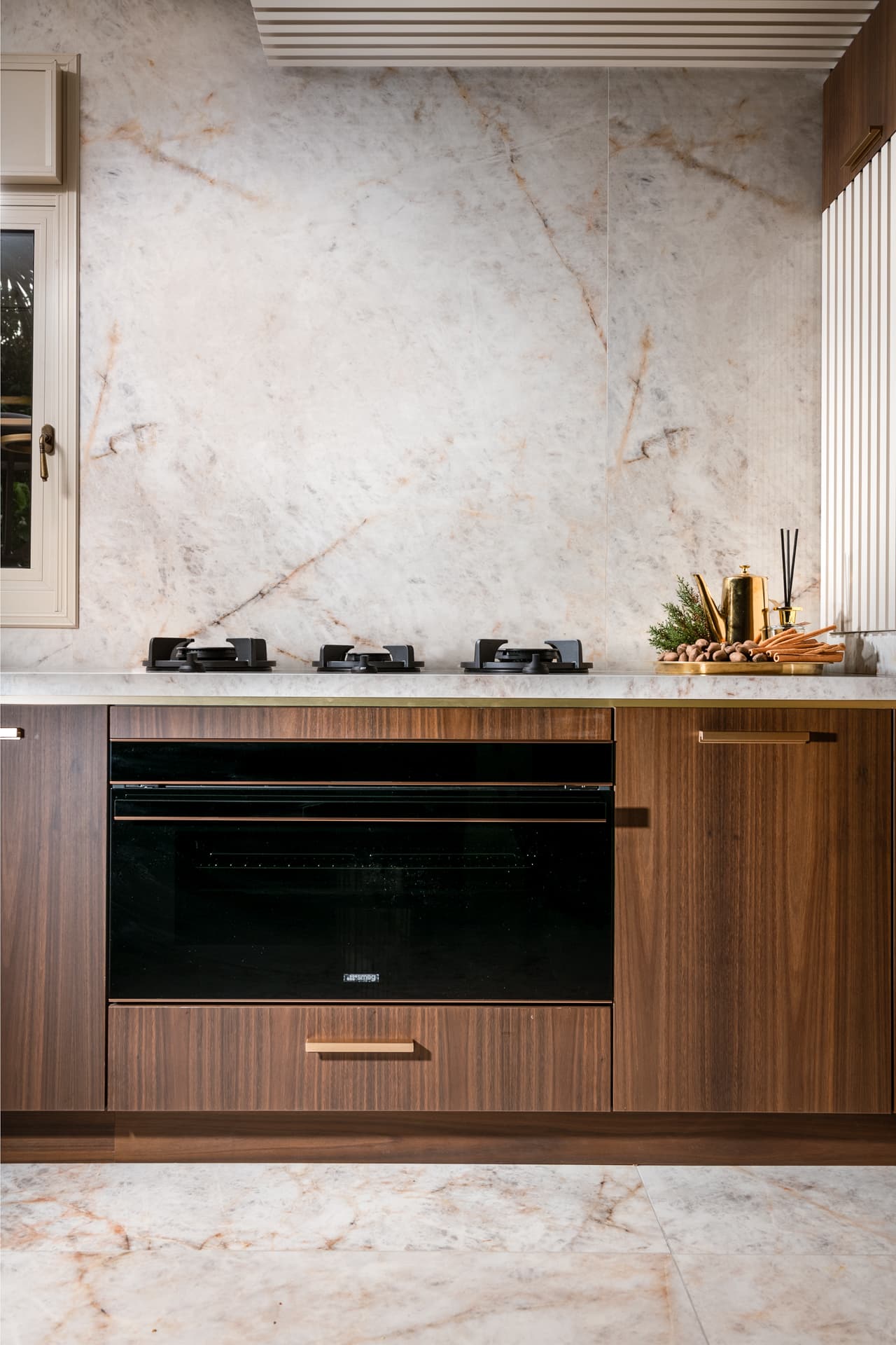 Walnut base cabinets with slim brass pulls framing a black built-in oven below a veined stone backsplash in the kitchen of Cognac