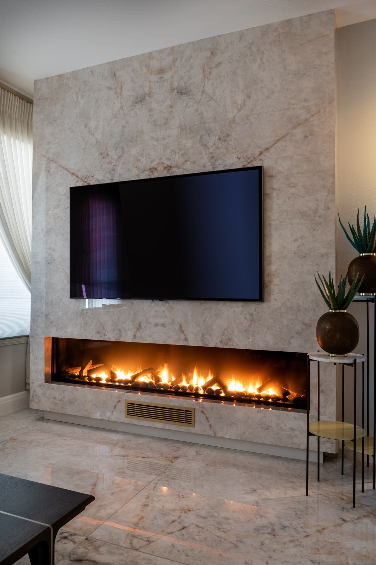 Veined stone fireplace wall with a long linear fire below a wall-mounted TV and a polished stone floor in the living room of Cognac