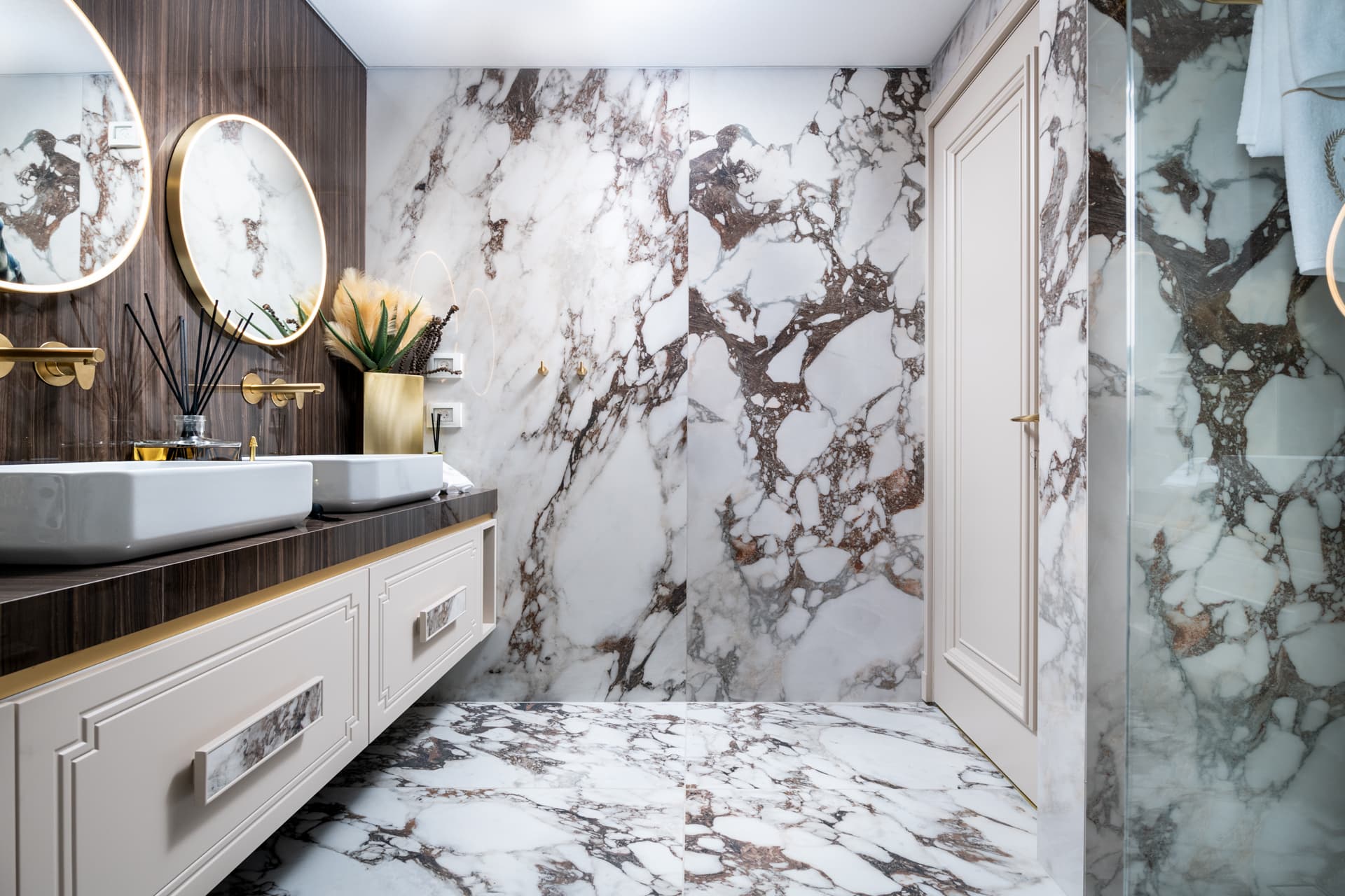 Bold dark-veined white stone walls and floor, a paneled door and the double vanity with round mirrors in the master bathroom of Cognac