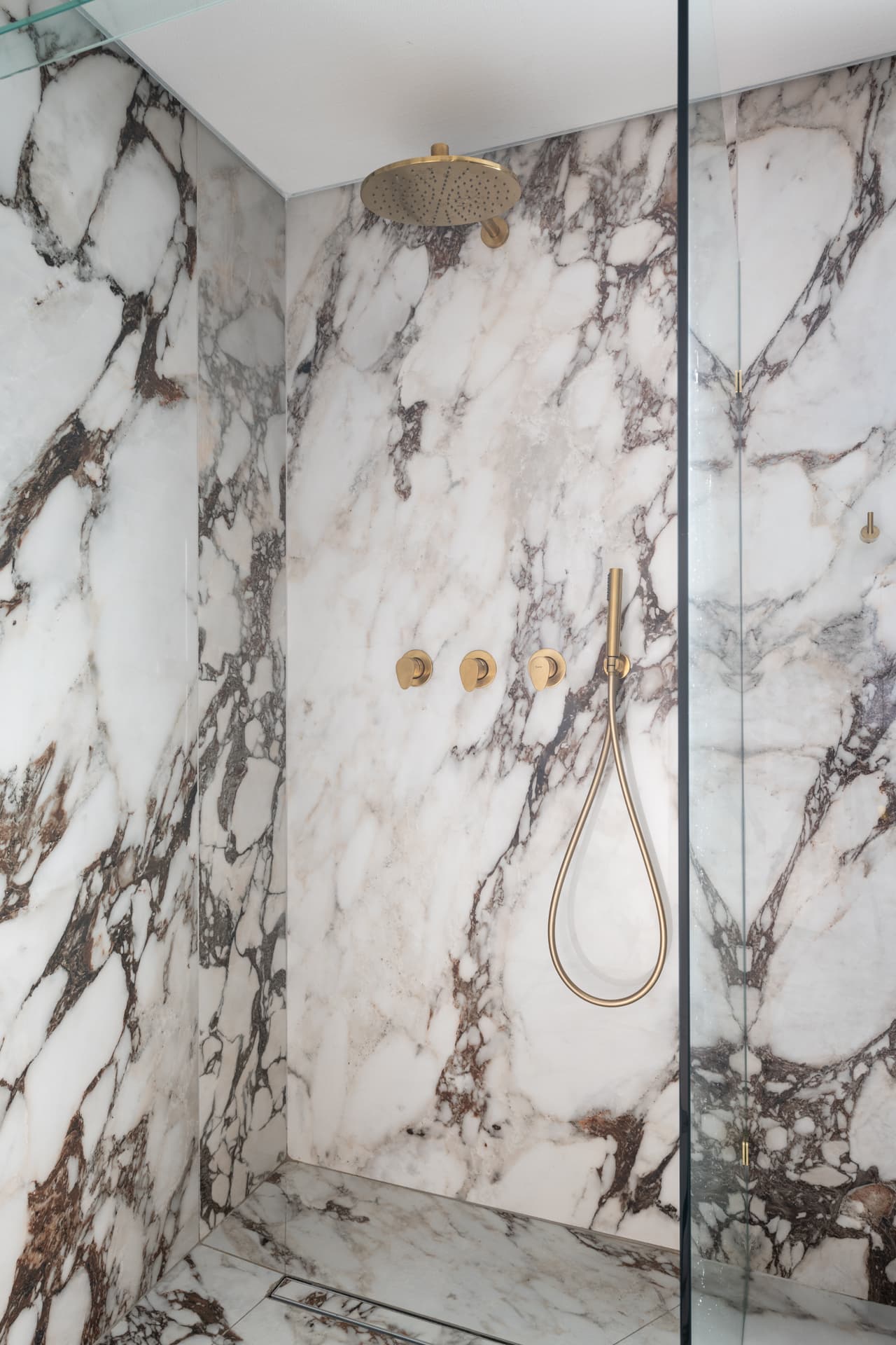 Glass-walled walk-in shower lined in dark-veined white stone with brushed brass rain shower and hand shower in the master bathroom of Cognac