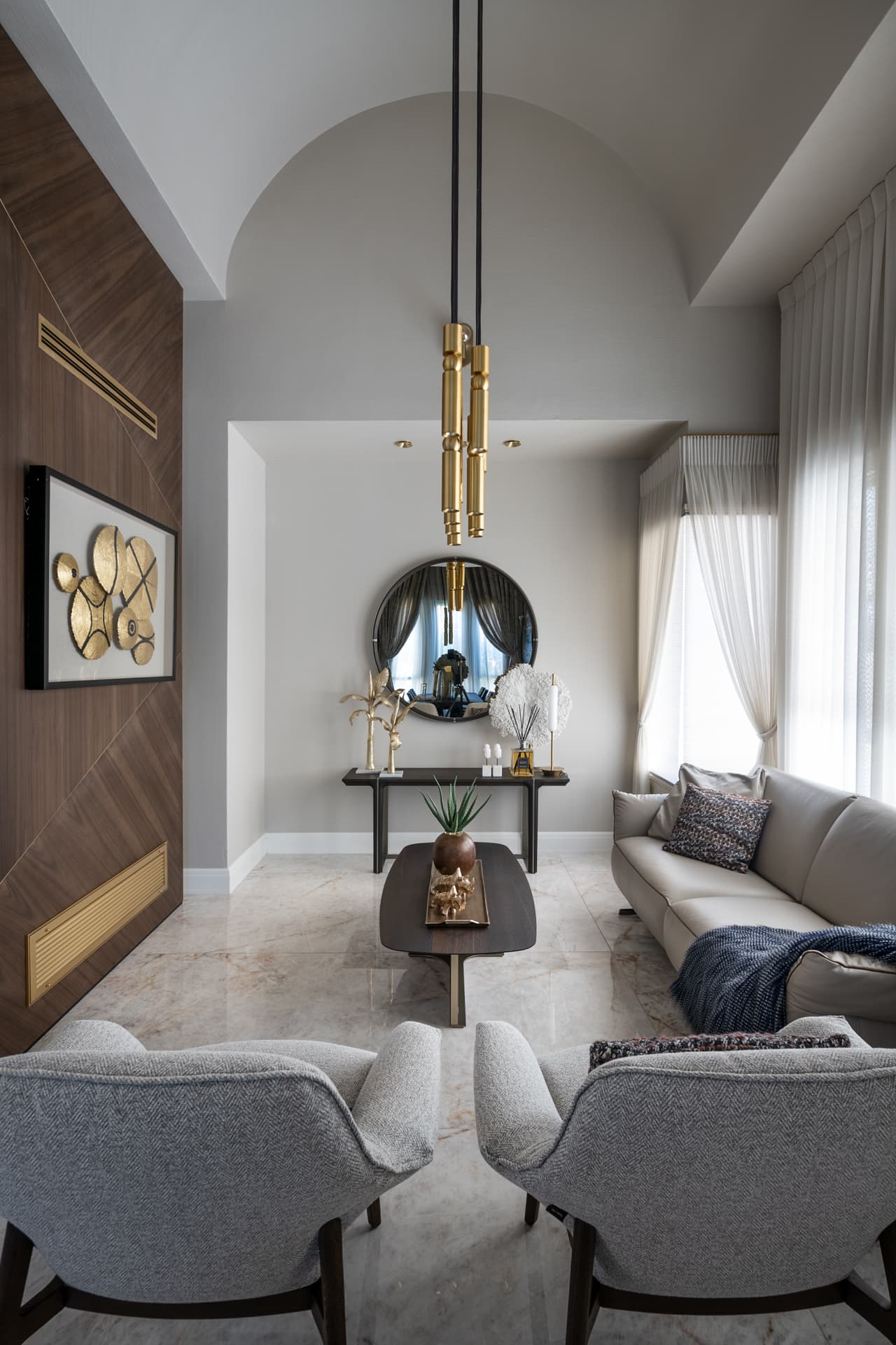 Daylit view of the vaulted salon with a brass tube pendant, a round mirror above a dark console and a beige sofa by sheer curtains in Cognac