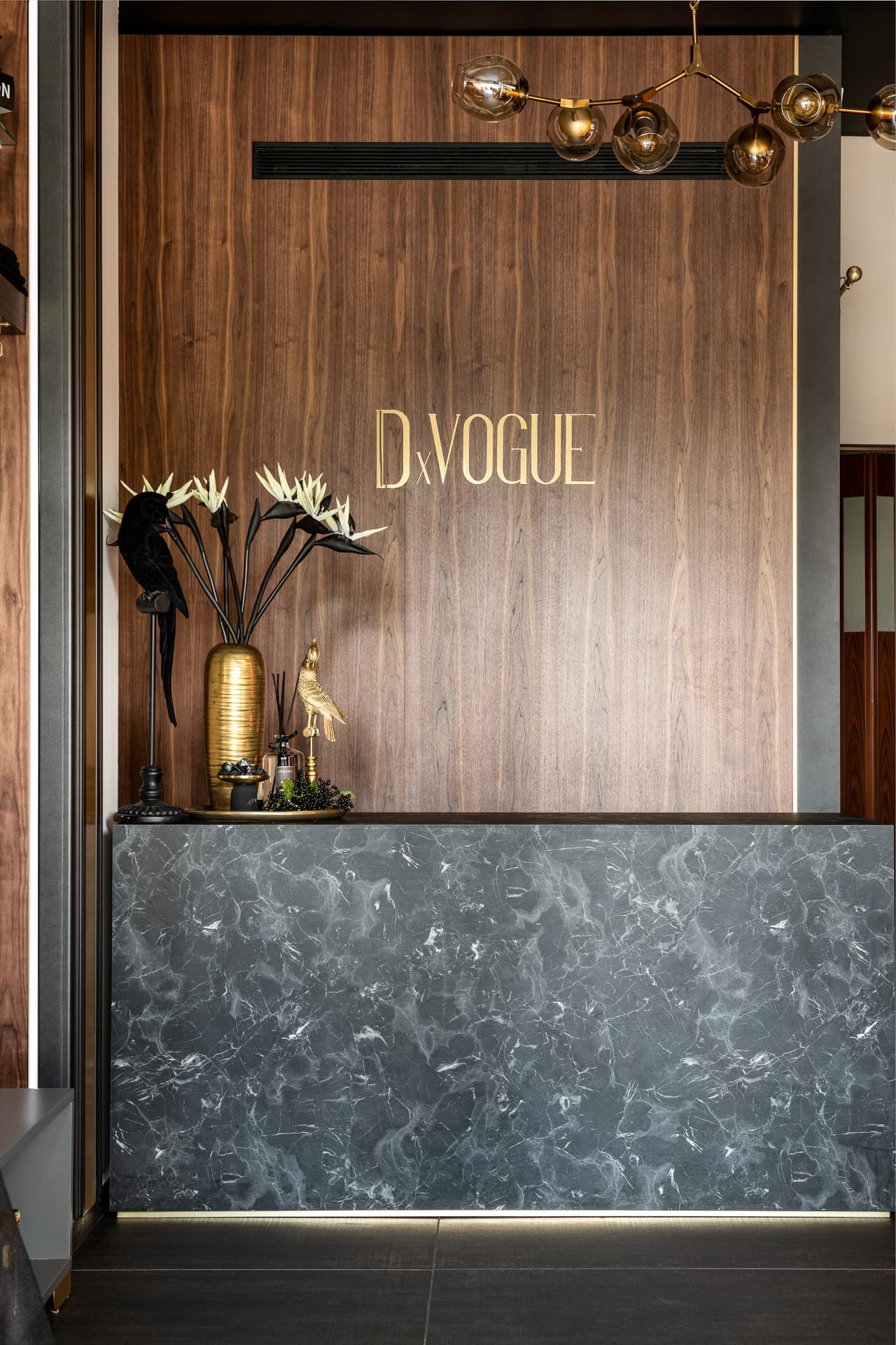 Dark veined stone counter with a lit base against a walnut-panelled wall under a brass globe chandelier, at the counter of DxVogue
